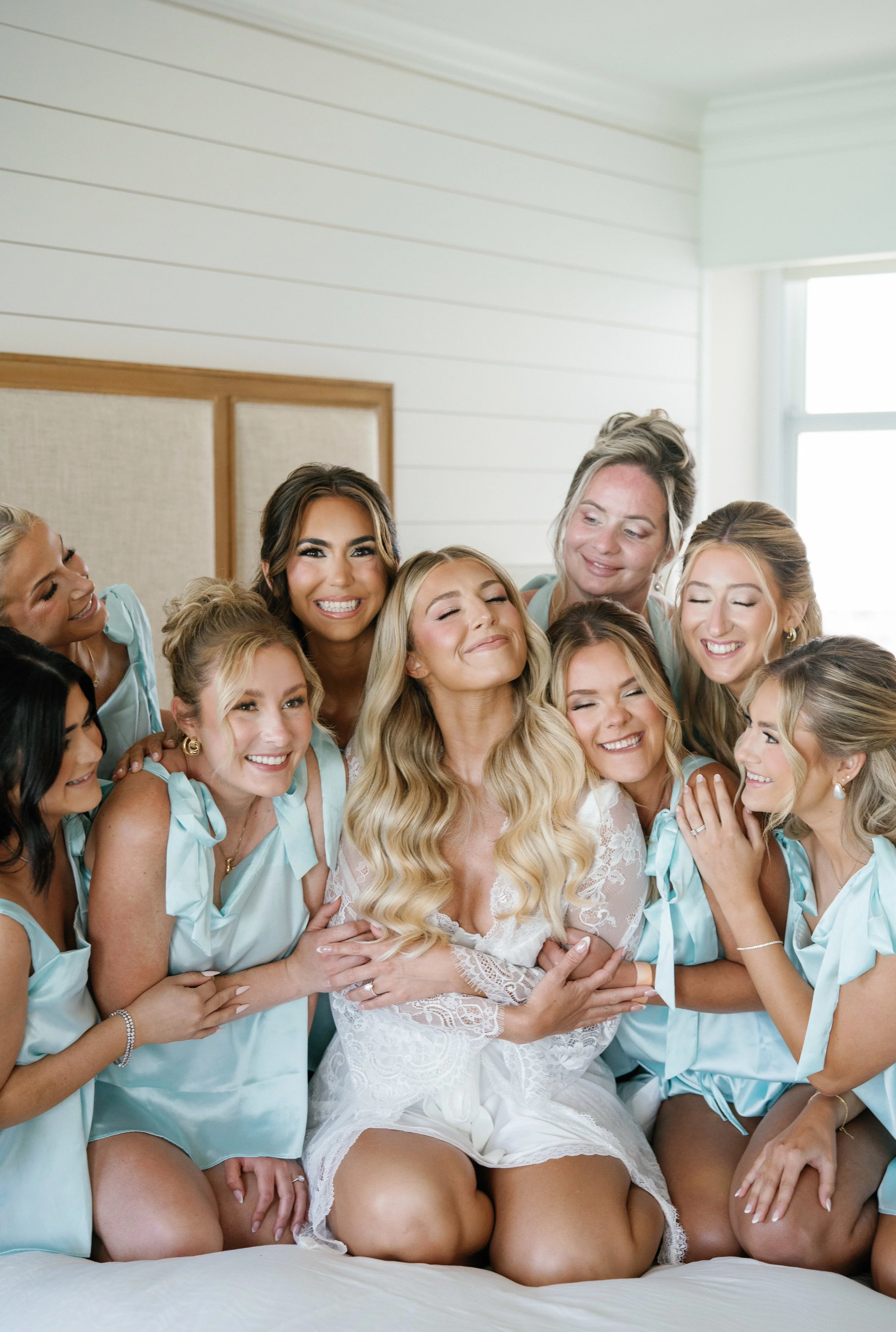 A group of women, likely bridesmaids, hugging a smiling bride with blonde hair in a white dress. The women are wearing matching light blue satin robes. The scene is indoors with white walls and natural light coming from a window.