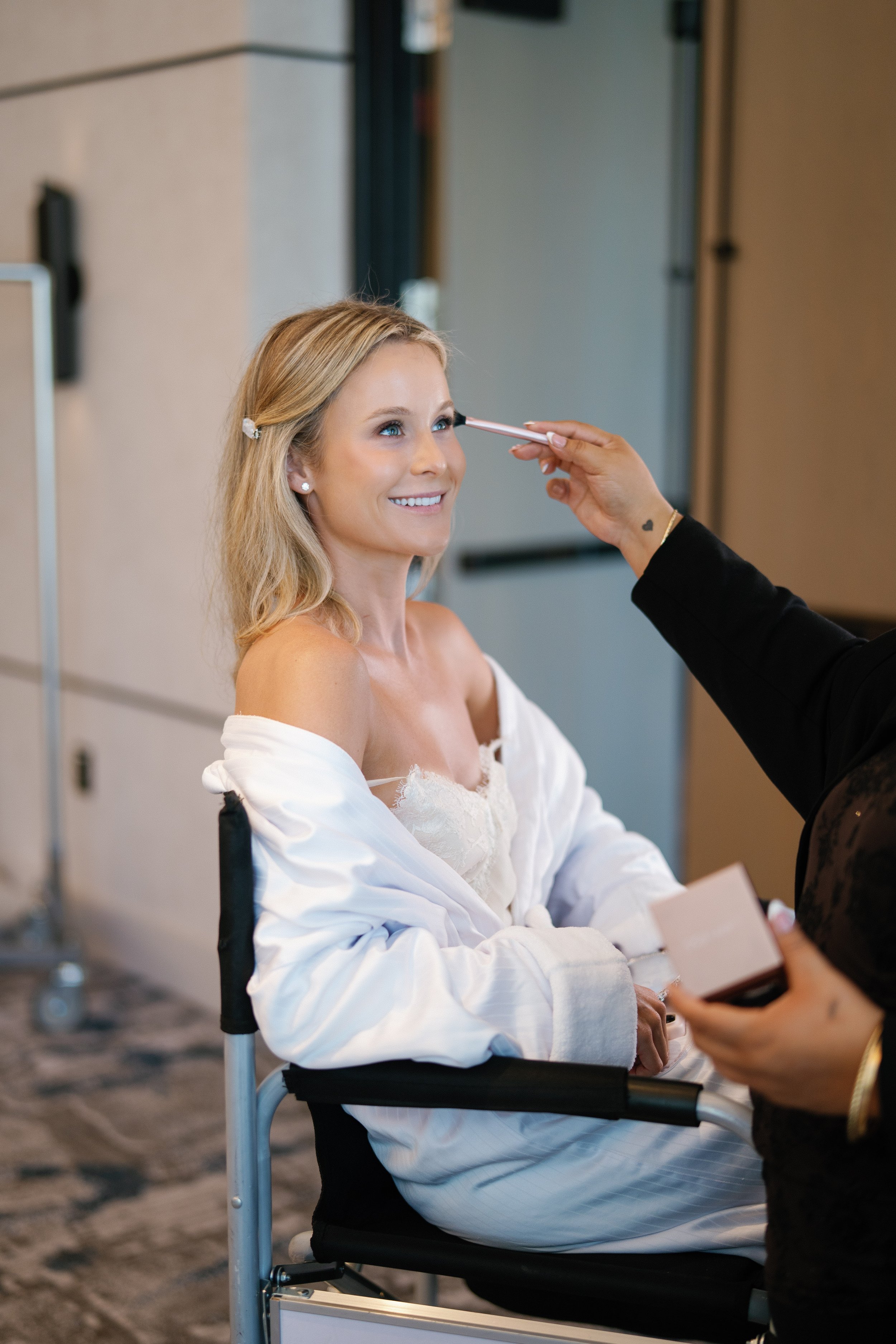 A smiling woman with blonde hair, sitting in a wheelchair, getting makeup applied by a person holding a brush near her face.