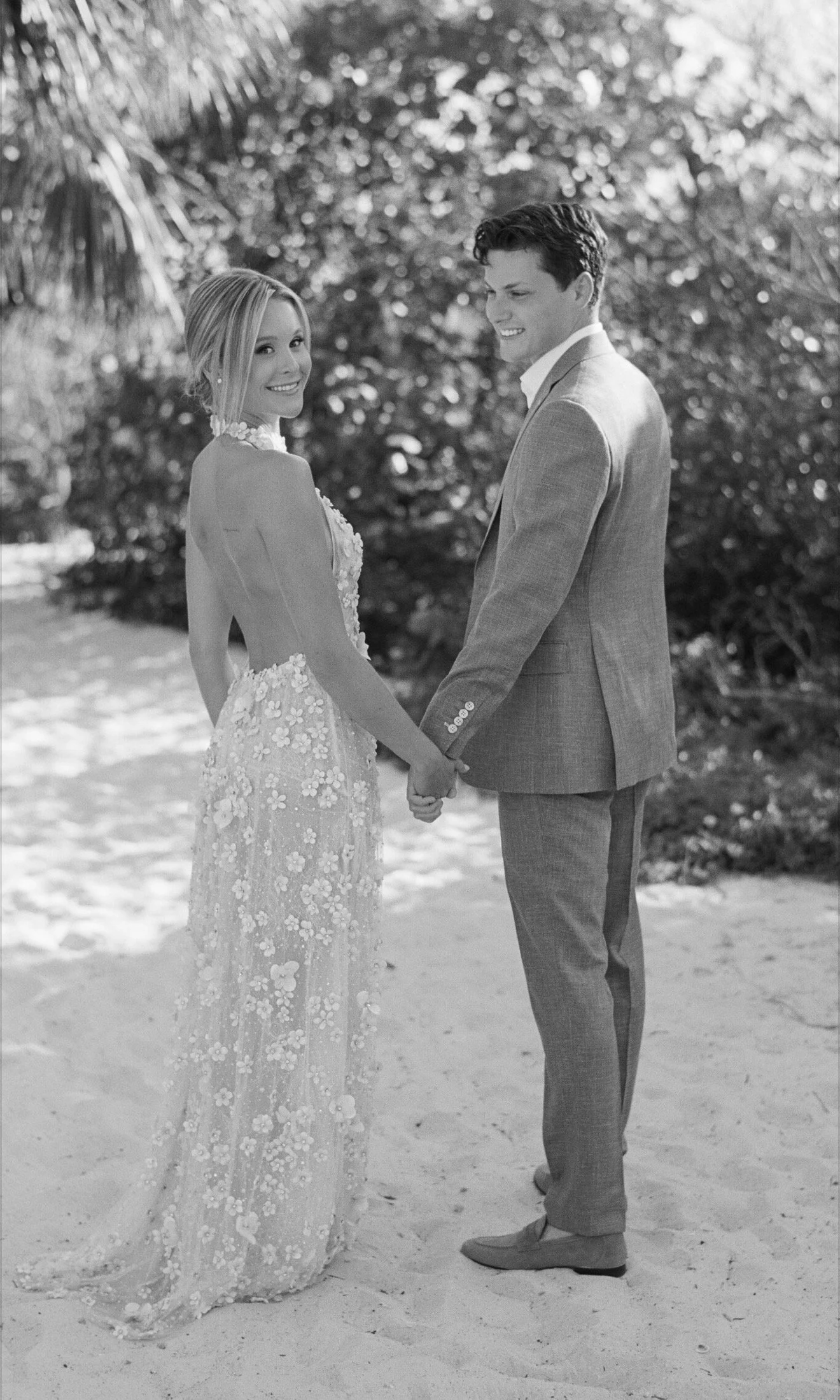 A black and white photo of a smiling woman in a floral dress and a man in a suit, holding hands and standing outdoors, with trees and sunlight in the background.