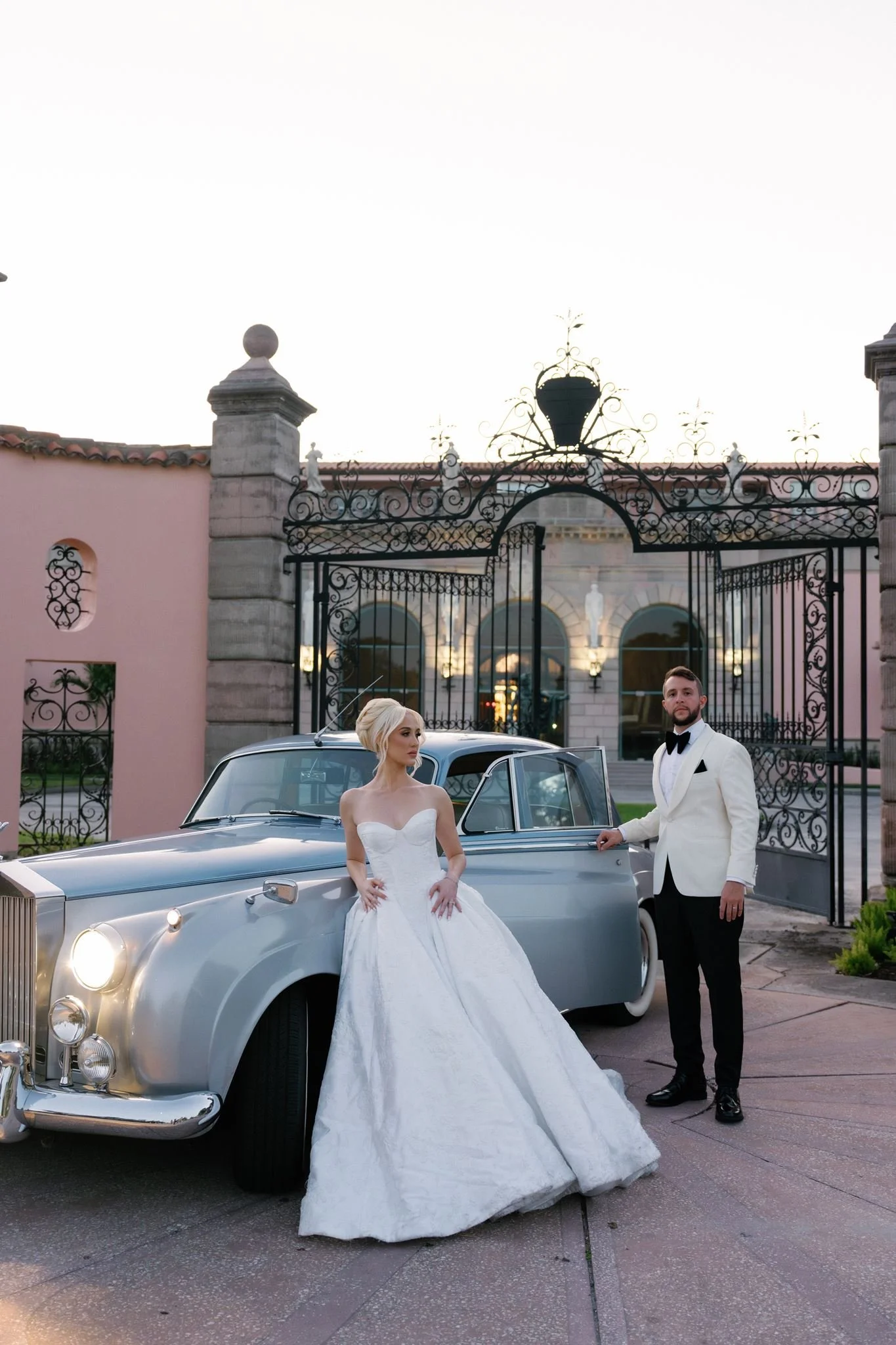 A bride in a white wedding gown leaning against a vintage silver car, and a groom in a tuxedo standing beside her, in front of an ornate wrought-iron gate and building with statues in the background during evening.