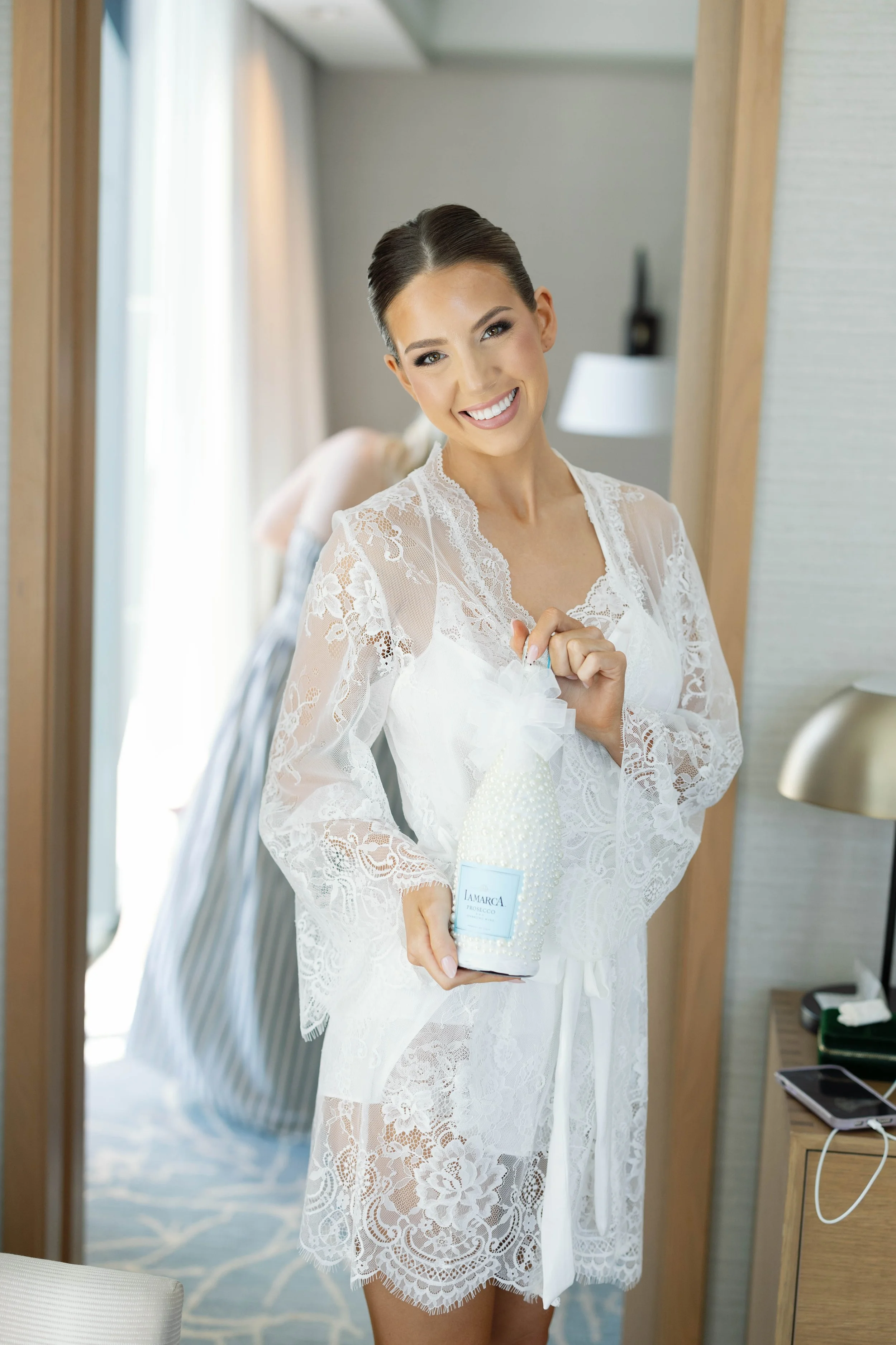 A woman in a white lace dress stands in front of a mirror, smiling and holding a bottle of La Marca Prosecco.