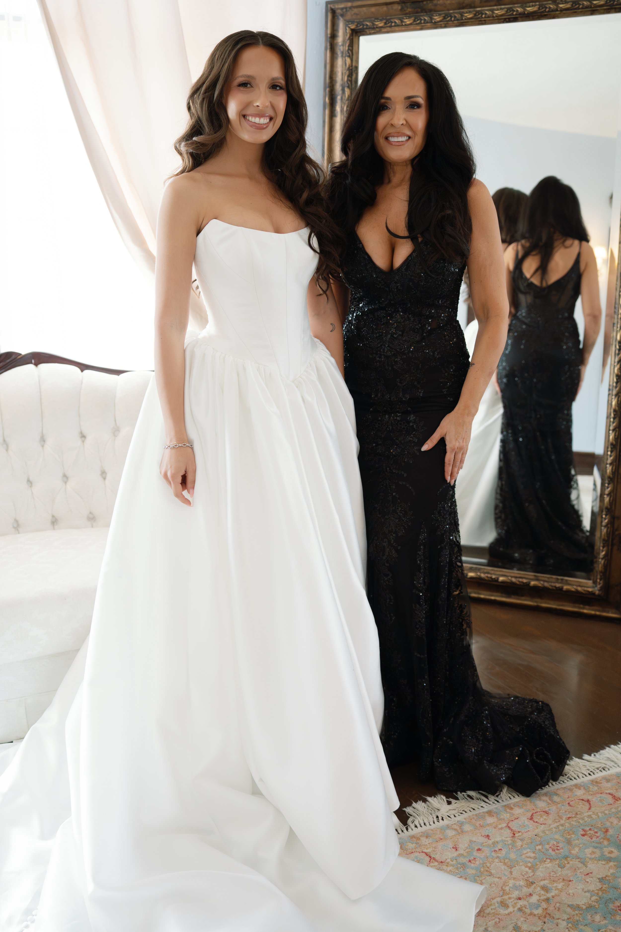 Two women standing indoors, one in a white wedding dress and the other in a black evening gown, smiling at the camera with a large mirror behind them.