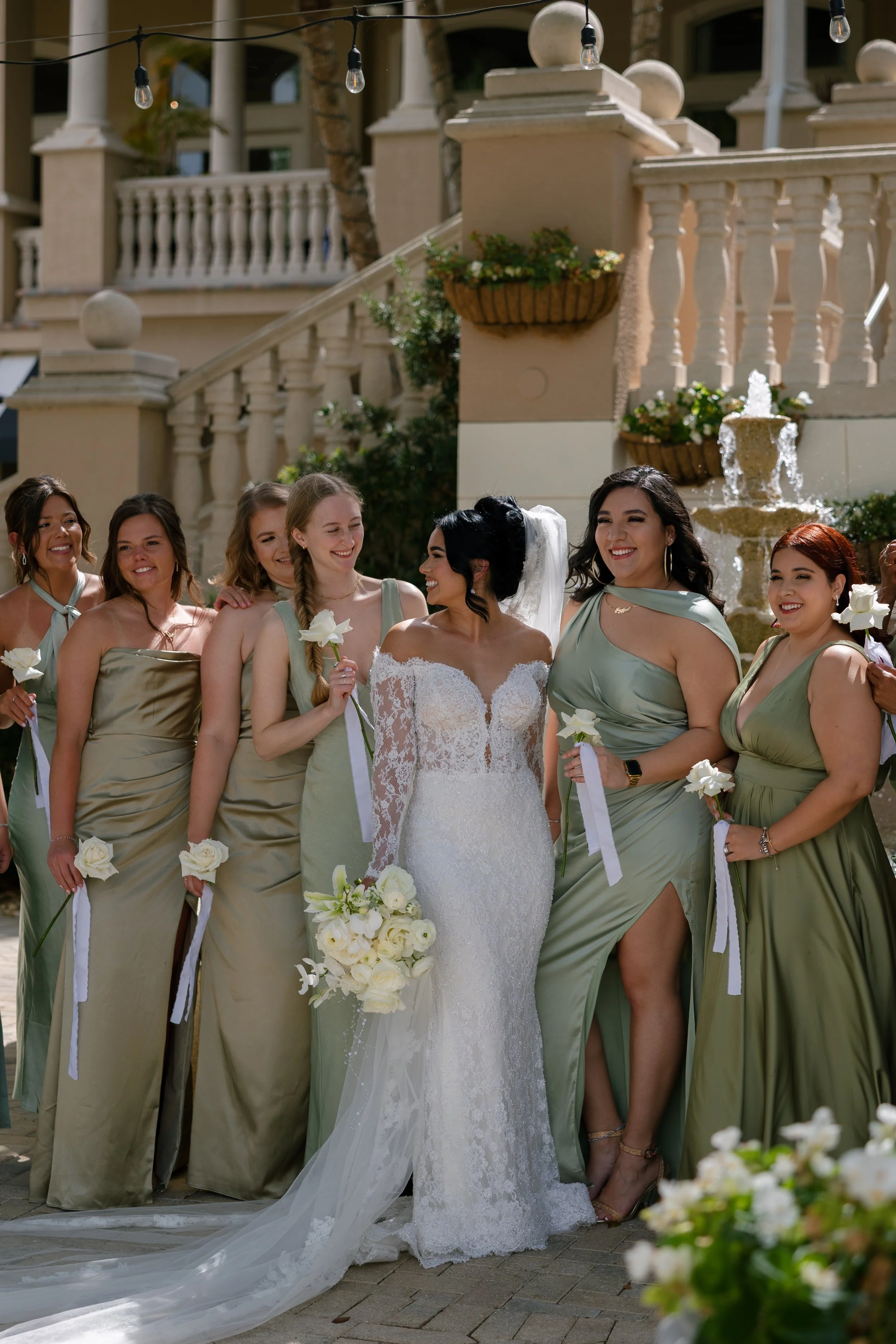 A group of women, including a bride in a white lace wedding gown holding a bouquet of white flowers, standing outside near a fountain and decorative balustrades. The women are dressed in matching sage green dresses and are smiling.