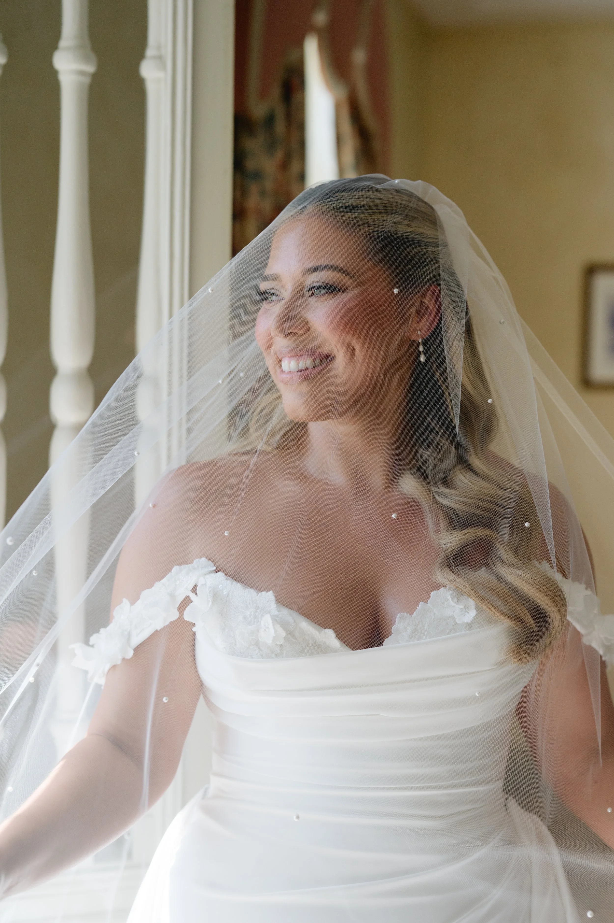 A bride with long blonde hair styled in loose waves, wearing a white lace wedding dress with off-the-shoulder sleeves, and a sheer veil with small pearls, smiling while looking out a window with white spindled railing in the background.