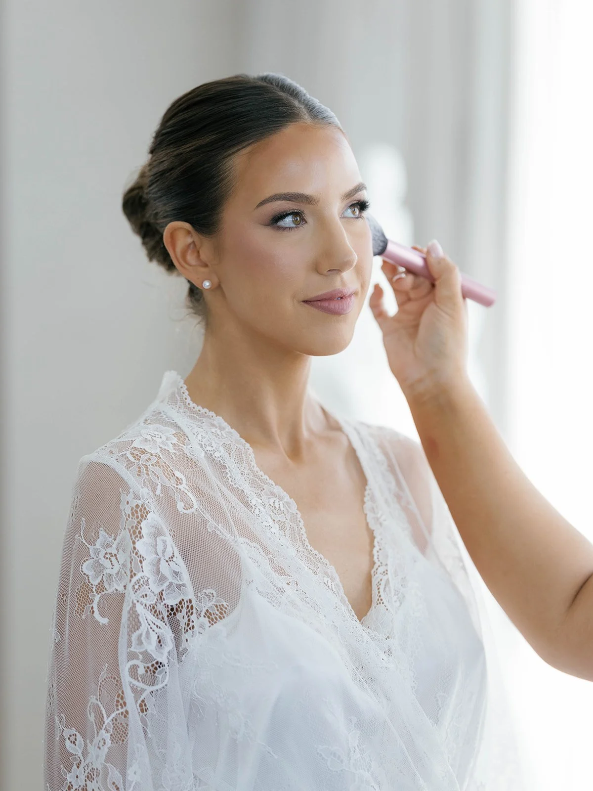 A woman with brown hair styled in an elegant updo and wearing pearl earrings is having her makeup done by a makeup artist using a makeup brush. She is wearing a white lace dress with floral patterns.