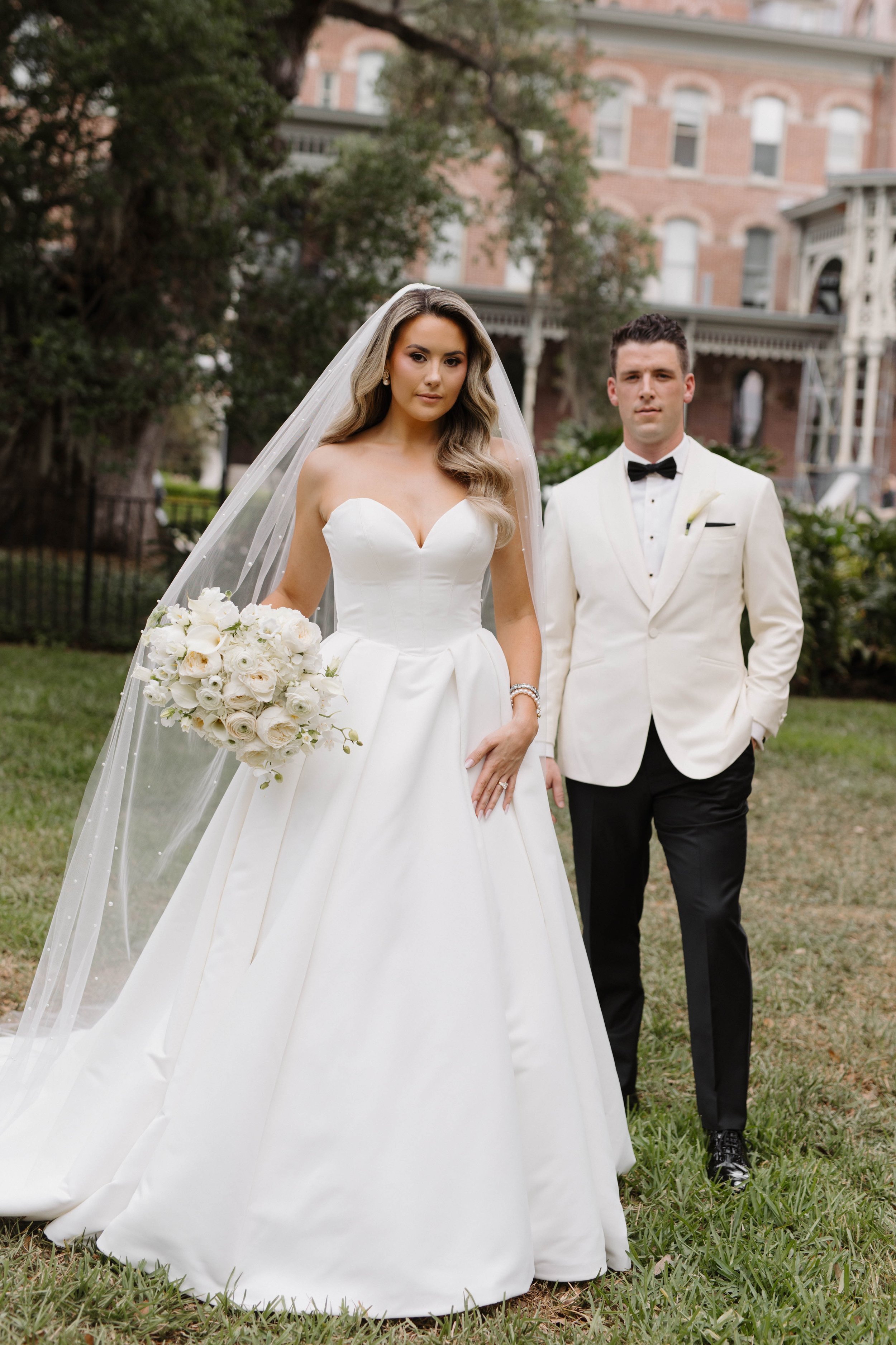 Bride in a white strapless wedding dress with a veil holding a bouquet of white roses and flowers, standing beside groom in a white tuxedo with black bow tie, in front of a historic brick building with greenery  Oxford exchange in Tampa fl . 
