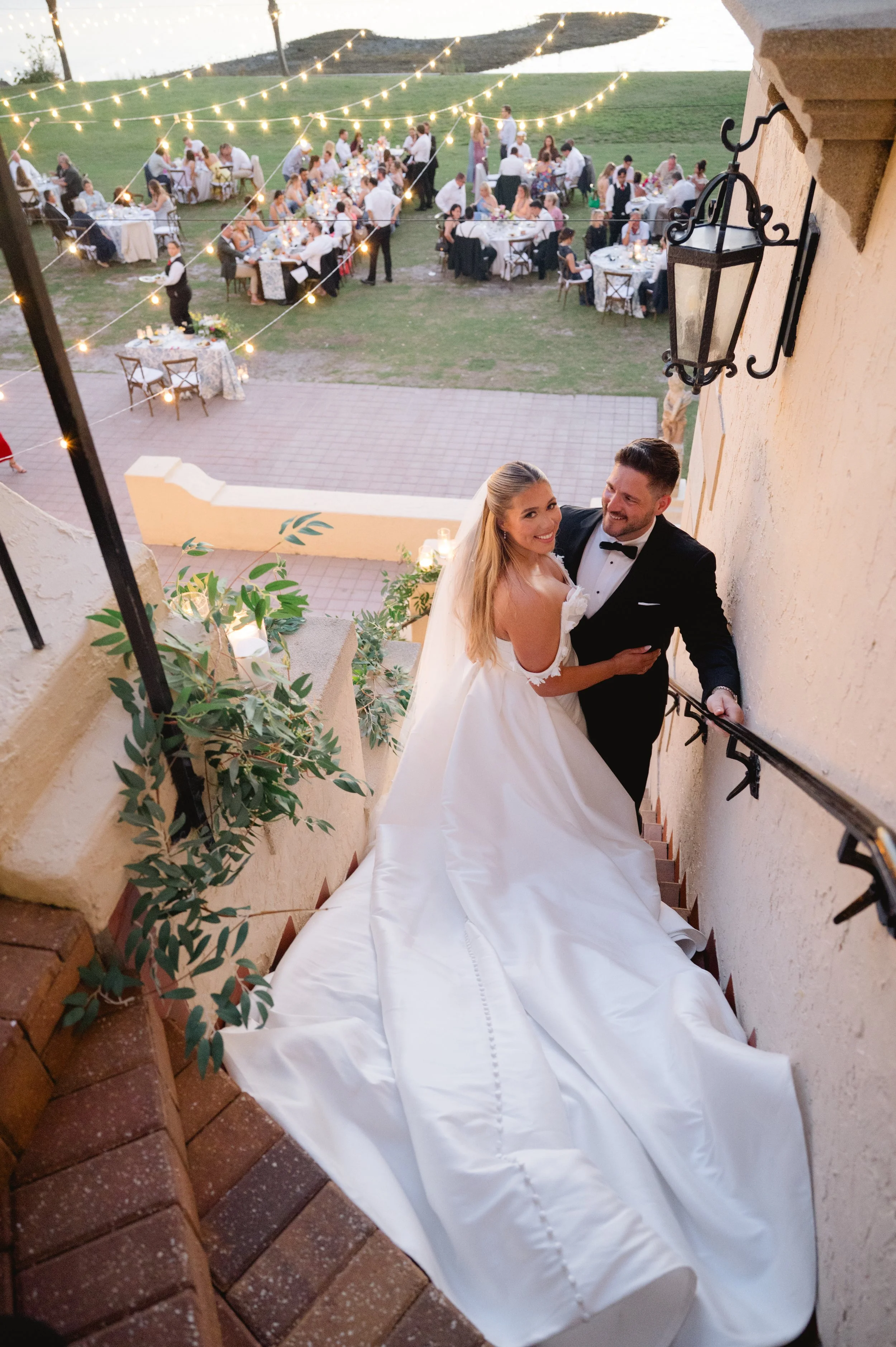 A newlywed couple smiling at each other on a staircase at their outdoor wedding reception, with tables and guests in the background under string lights at dusk.