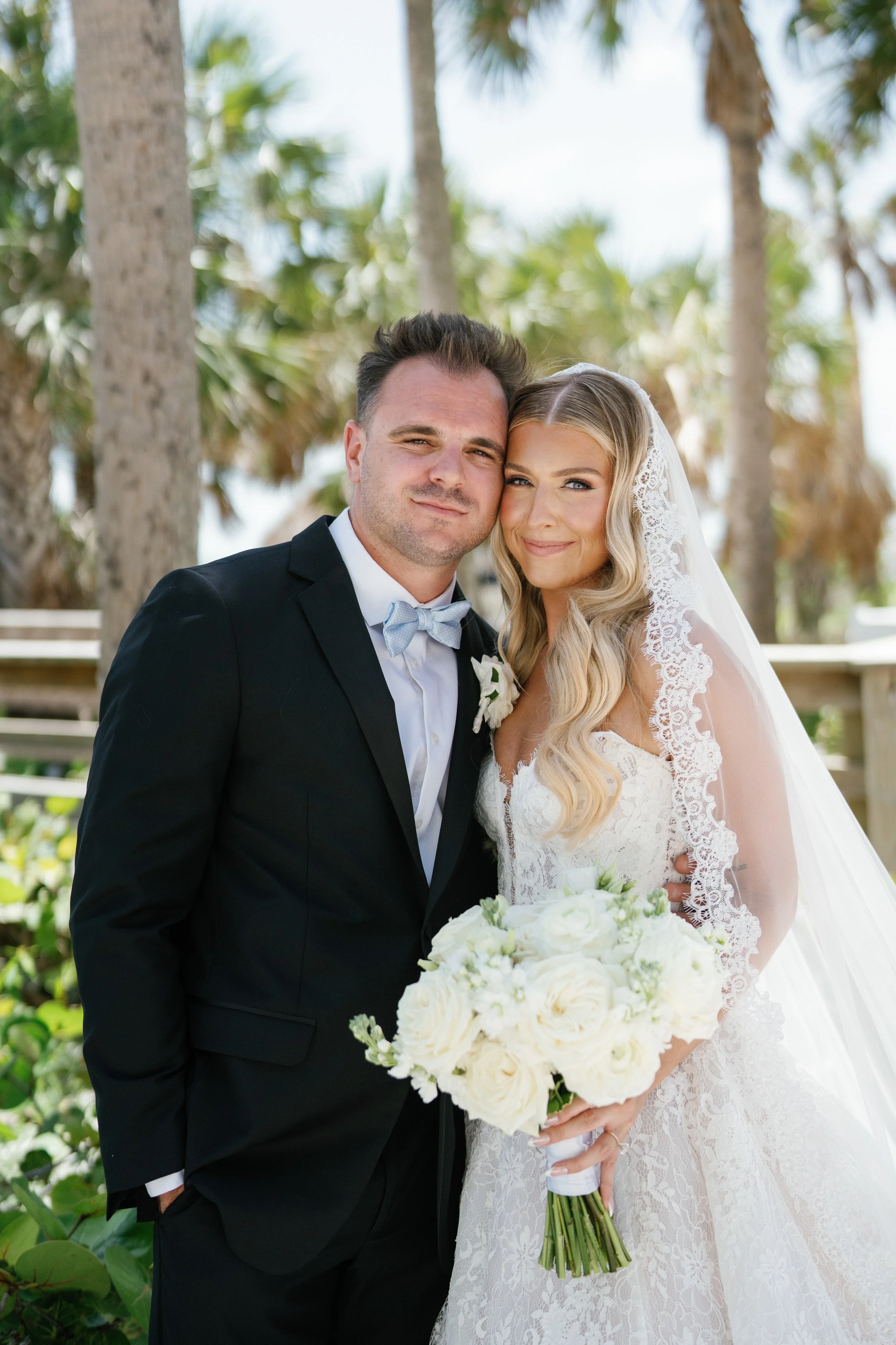 A newlywed couple stands close together outdoors among trees, with the groom in a black suit and light blue bow tie, and the bride in a white lace wedding dress and veil, holding a bouquet of white flowers.