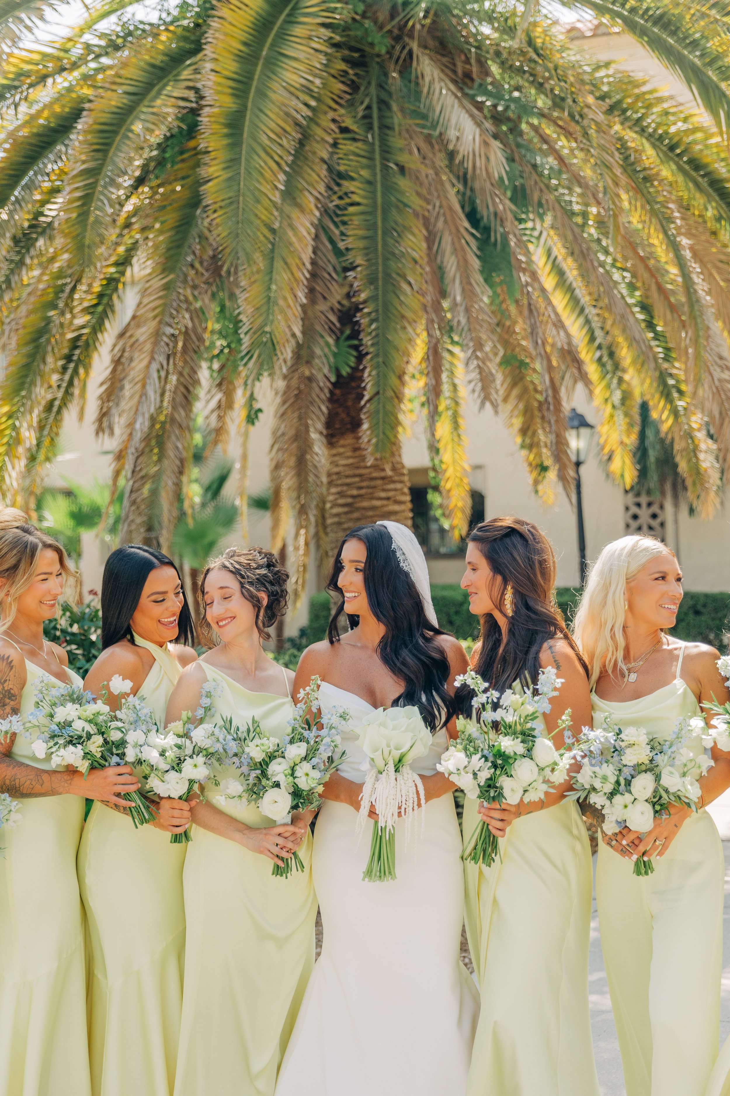 A group of women in yellow dresses holding bouquets of white flowers, standing outdoors beneath a large palm tree, celebrating a wedding.
