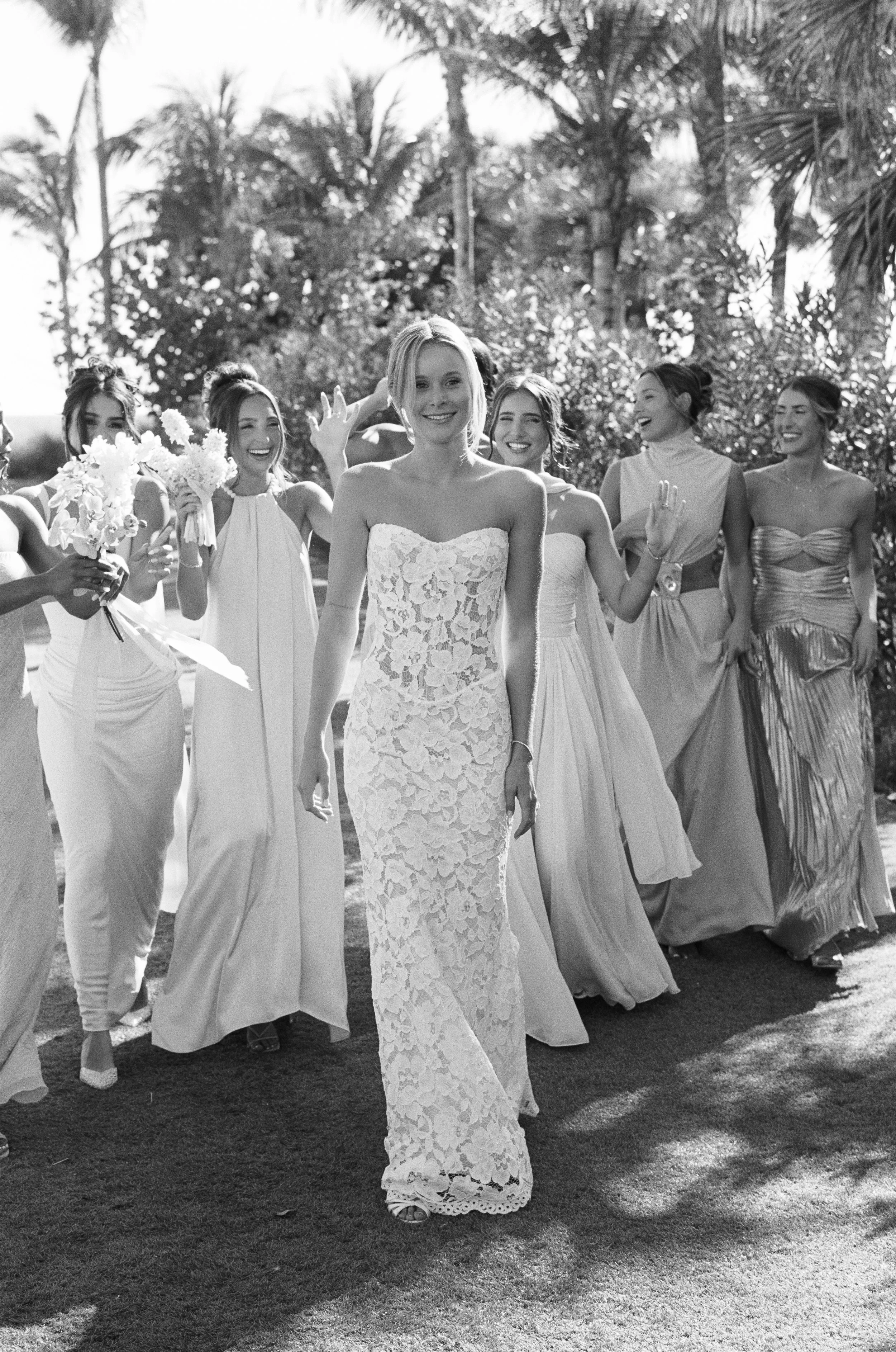 A bride in a strapless lace wedding dress walking outdoors with bridesmaids in light-colored dresses, surrounded by palm trees and foliage, sunny weather, black and white photograph.