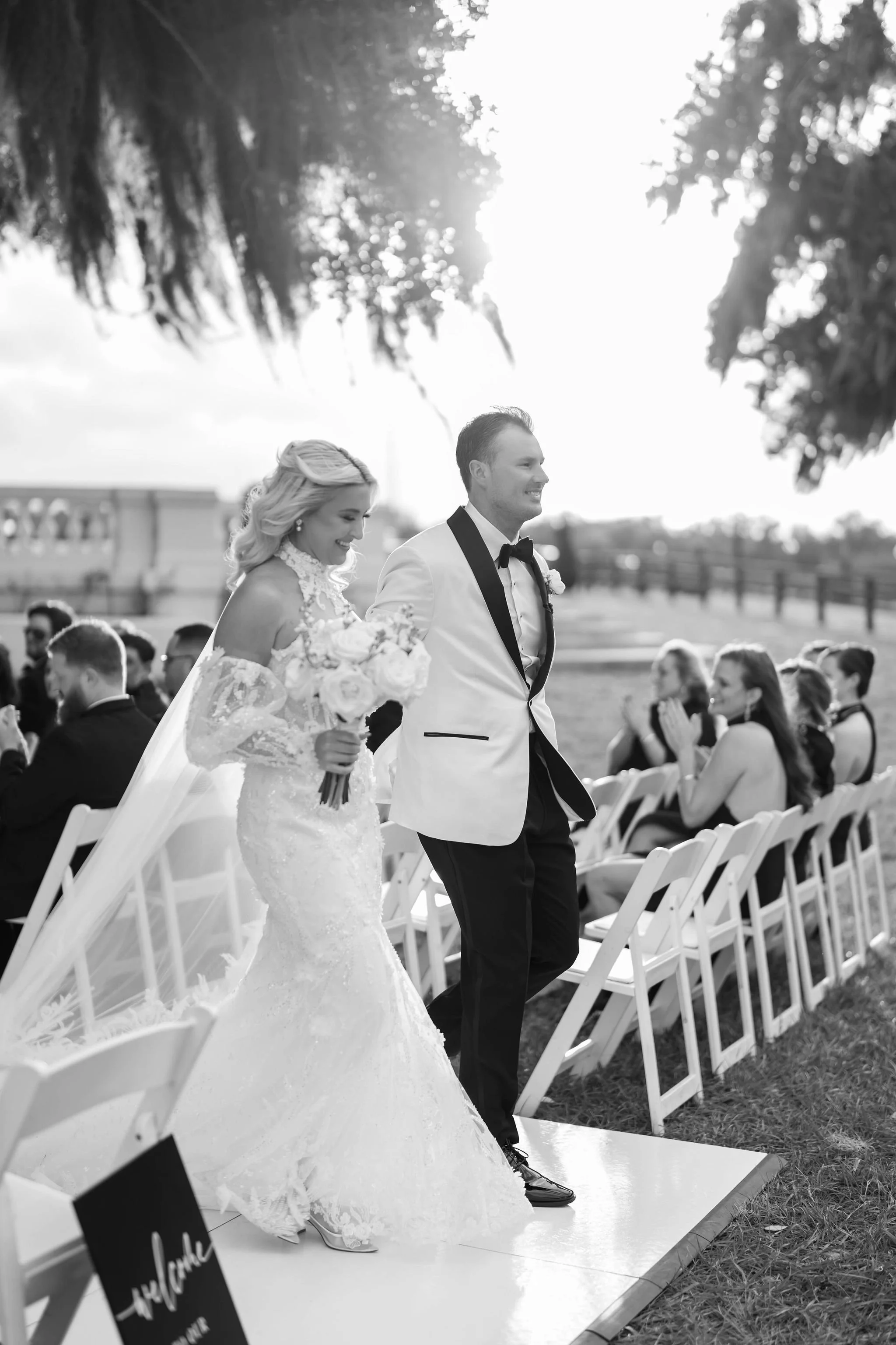 A bride and groom walking down the aisle during their wedding ceremony outdoors, with guests seated on either side, in a bright setting with trees in the background.