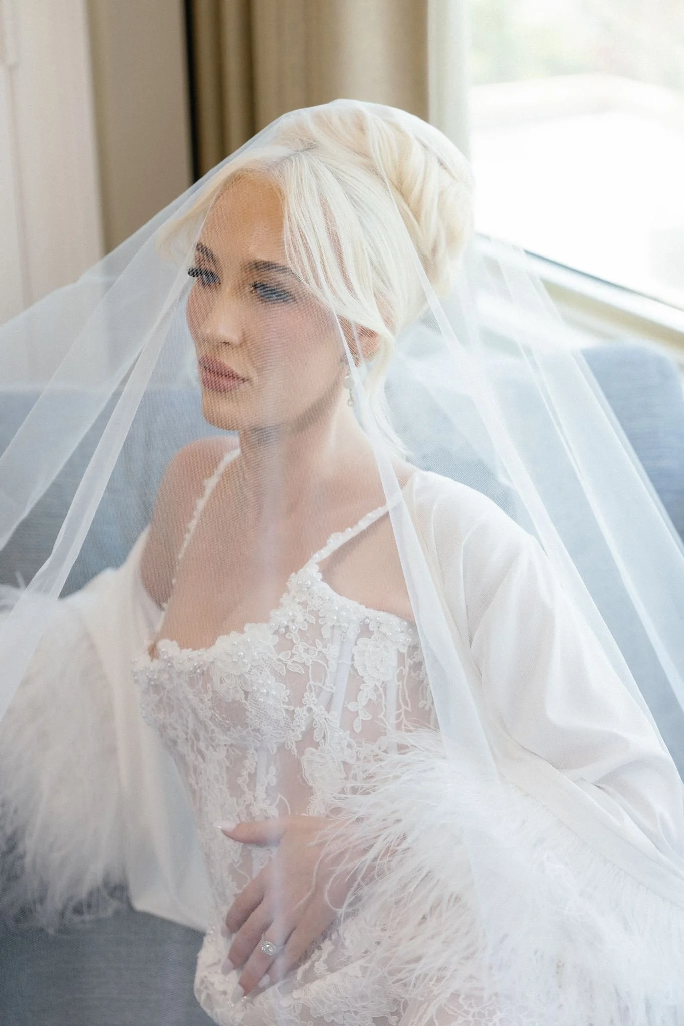 A bride with platinum blonde hair wearing a lace wedding gown with lace, pearls, and embroidery, a veil, and a diamond ring on her left hand.
