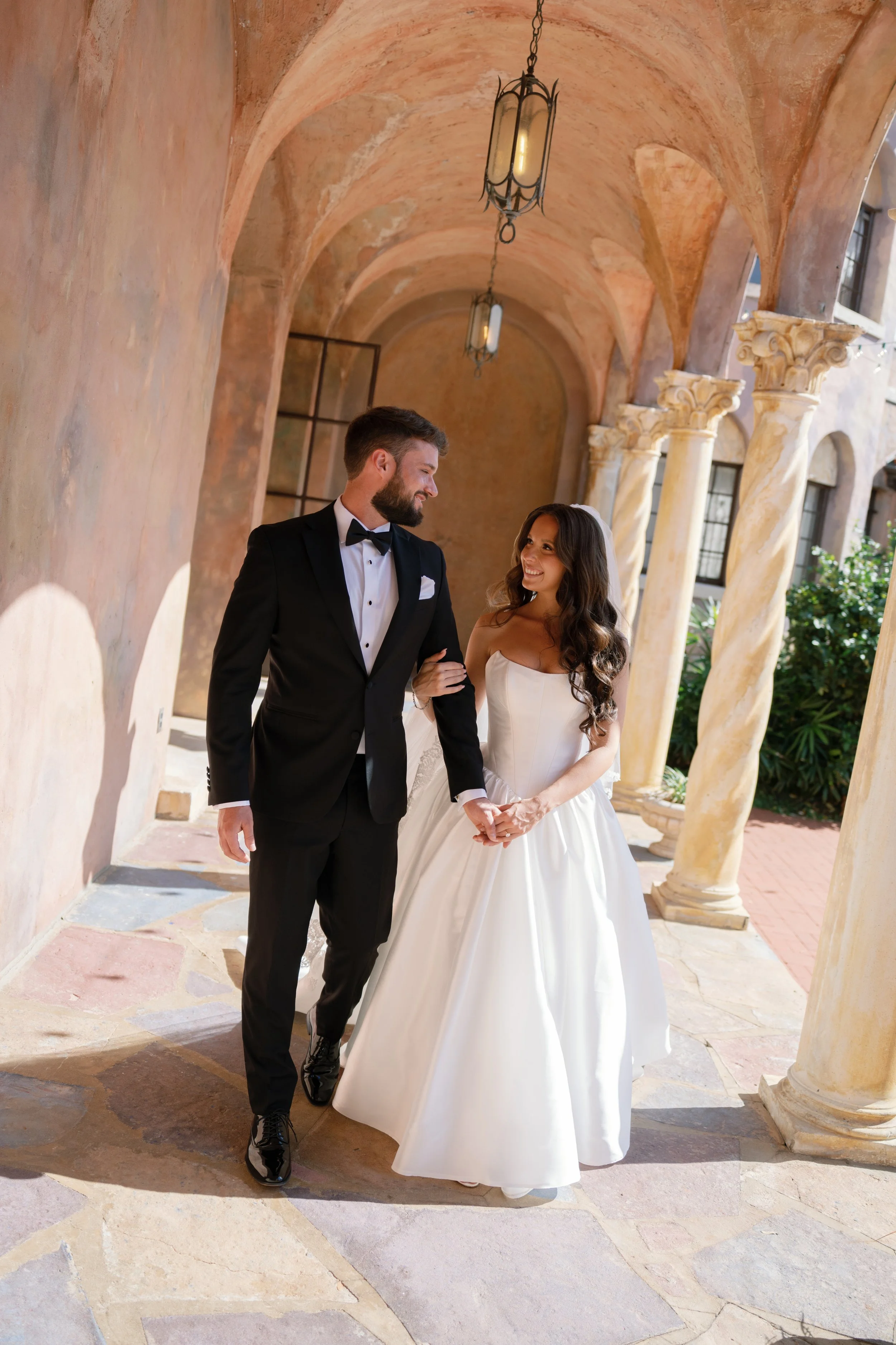A bride and groom walking arm in arm in a courtyard with tall arches and columns, the groom is wearing a black tuxedo and the bride a white wedding gown. They are smiling at each other.