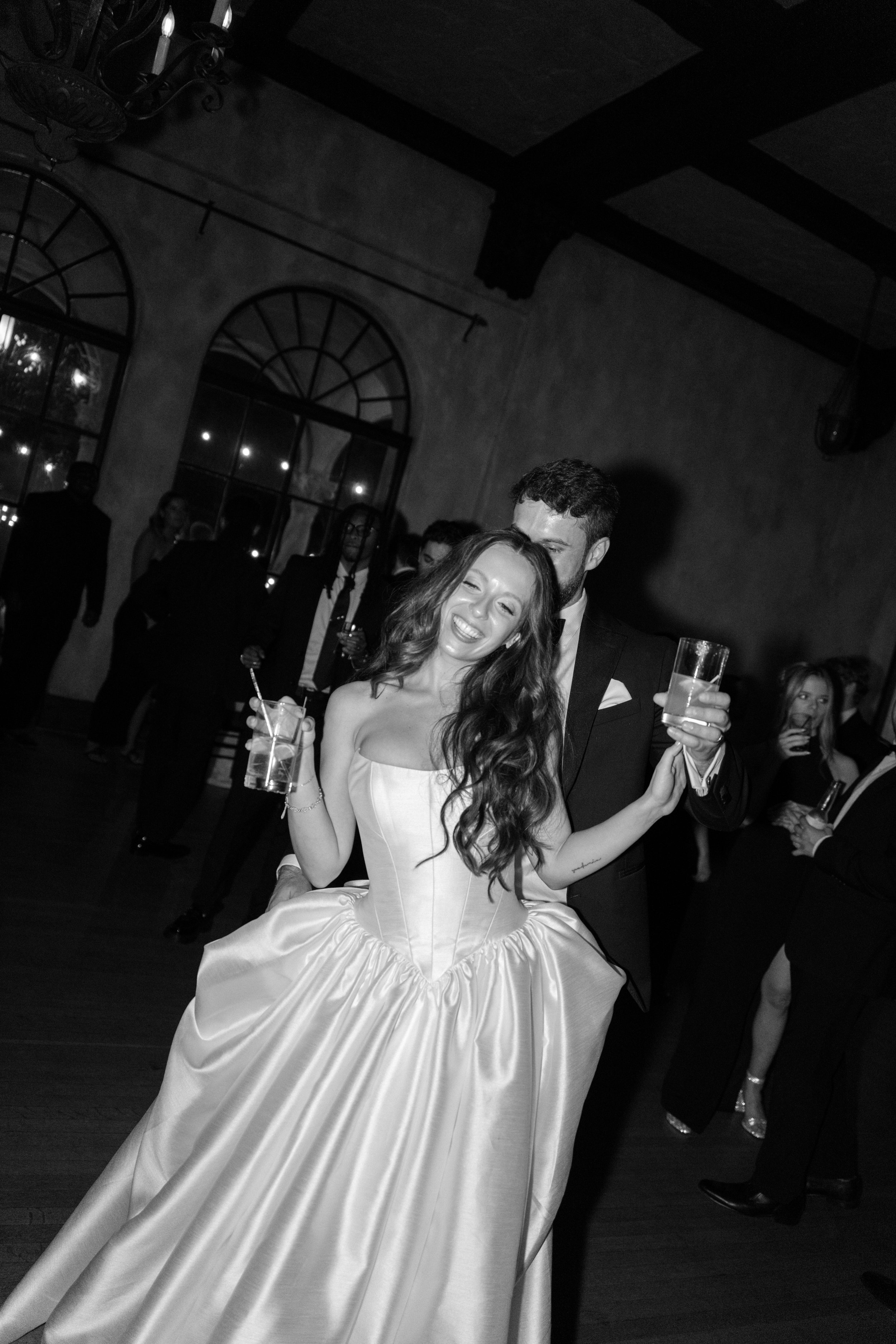 A bride and groom dancing at a wedding reception, with the bride wearing a strapless wedding dress and the groom wearing a black tuxedo, both holding drinks and smiling.
