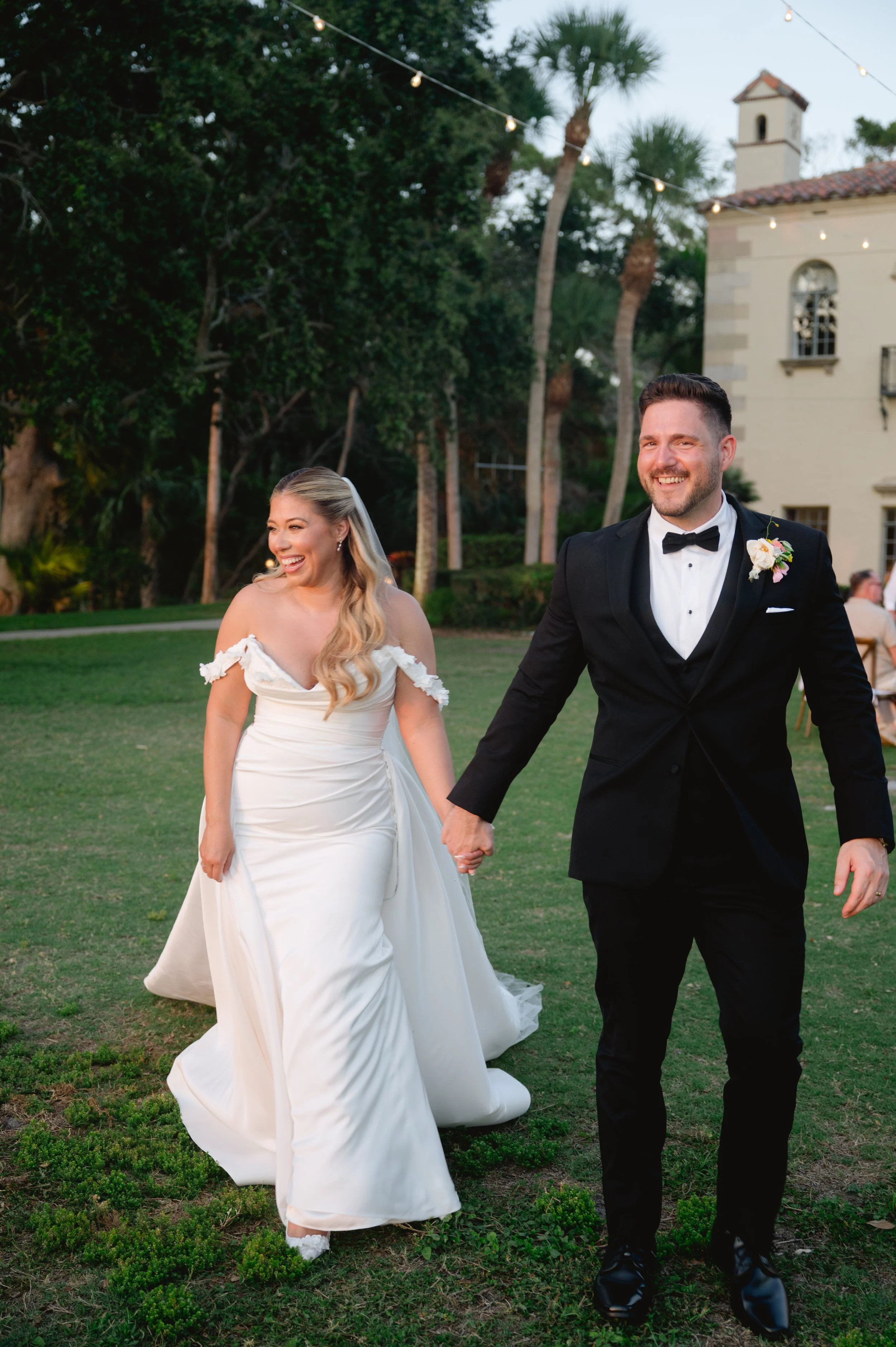 A bride and groom smiling and holding hands outdoors during a wedding celebration. The bride is wearing a white off-shoulder gown and the groom is dressed in a black tuxedo. There are trees, a building, and string lights in the background.