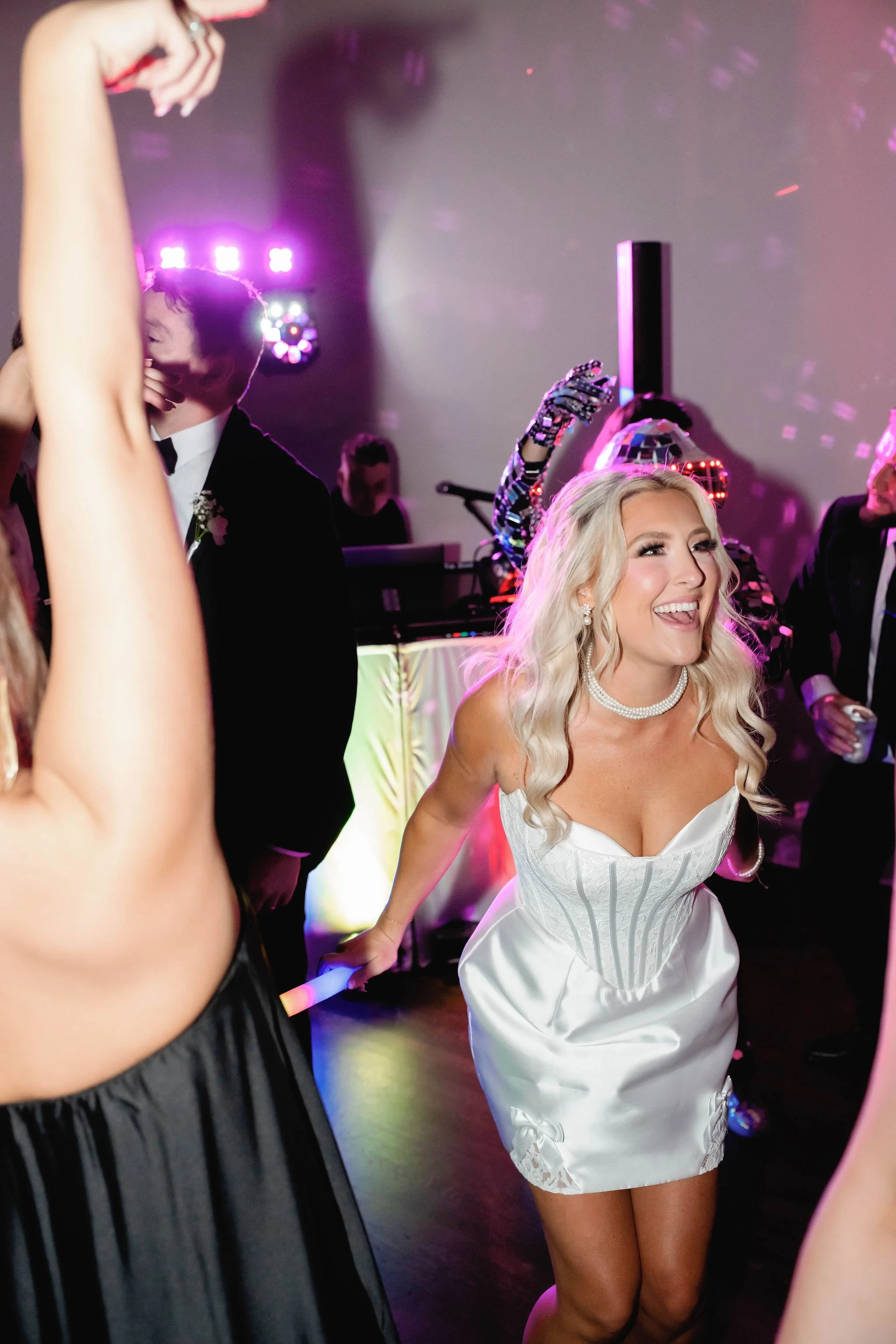 A woman in a white dress with corset details dancing at a party or celebration, smiling with blonde hair and jewelry, surrounded by other people and colorful lights.