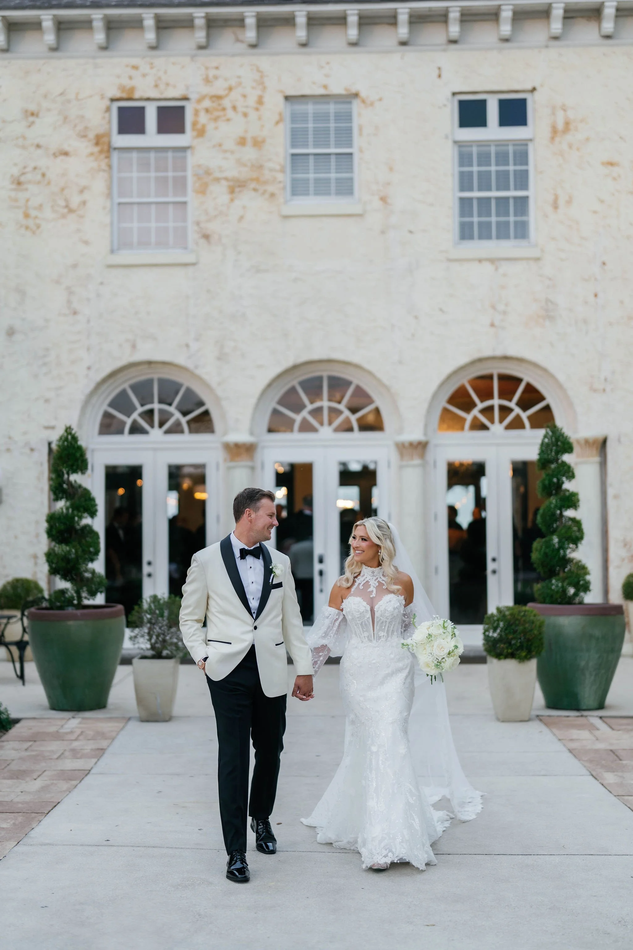 A bride and groom walking hand in hand outside a building with large arched windows. The groom is wearing a cream tuxedo with a black bow tie, and the bride is in a white lace wedding gown holding a bouquet of white flowers.
