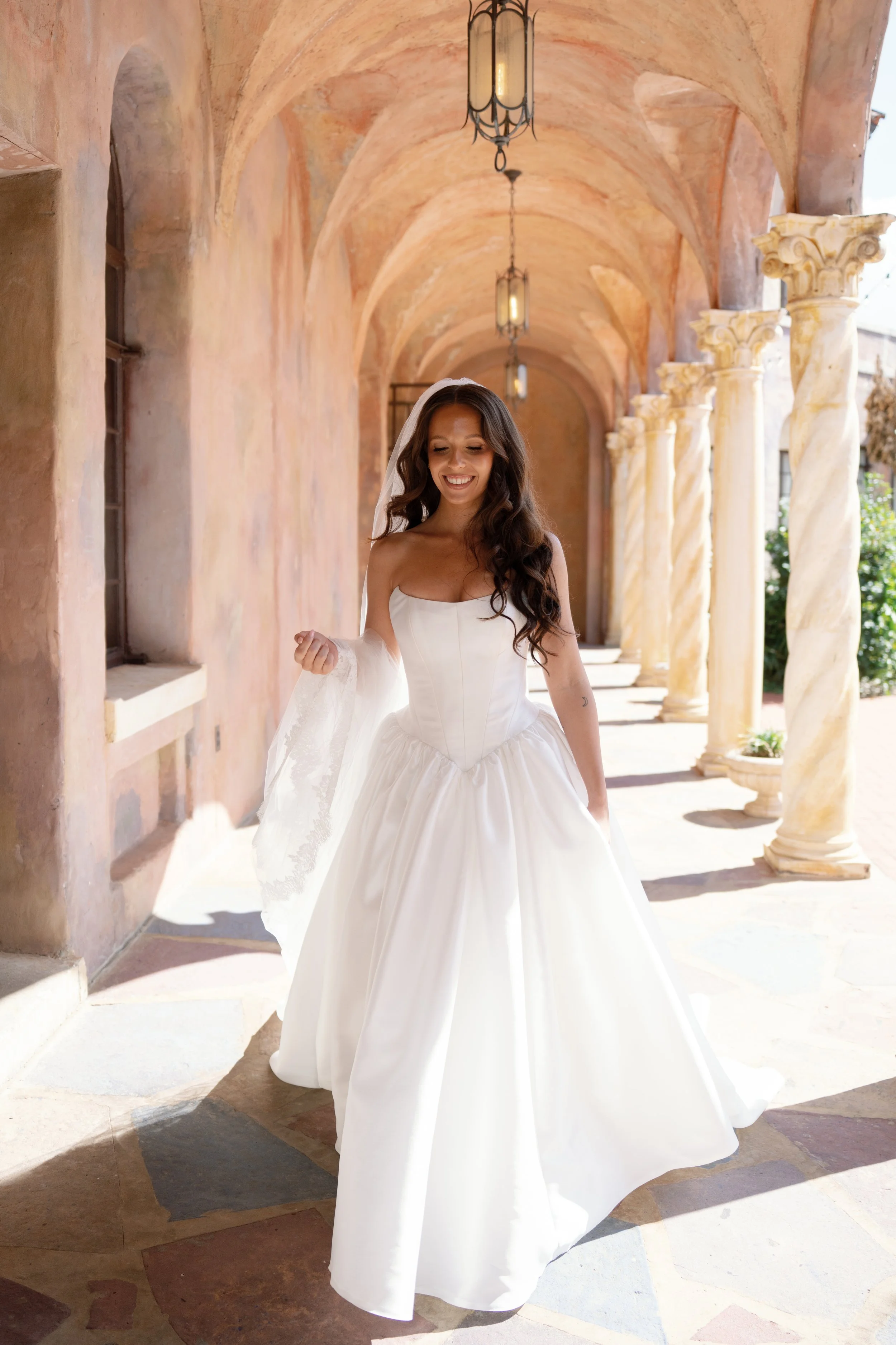 A woman in a white wedding gown smiling, standing under a covered walkway with arches and columns, bright sunlight.