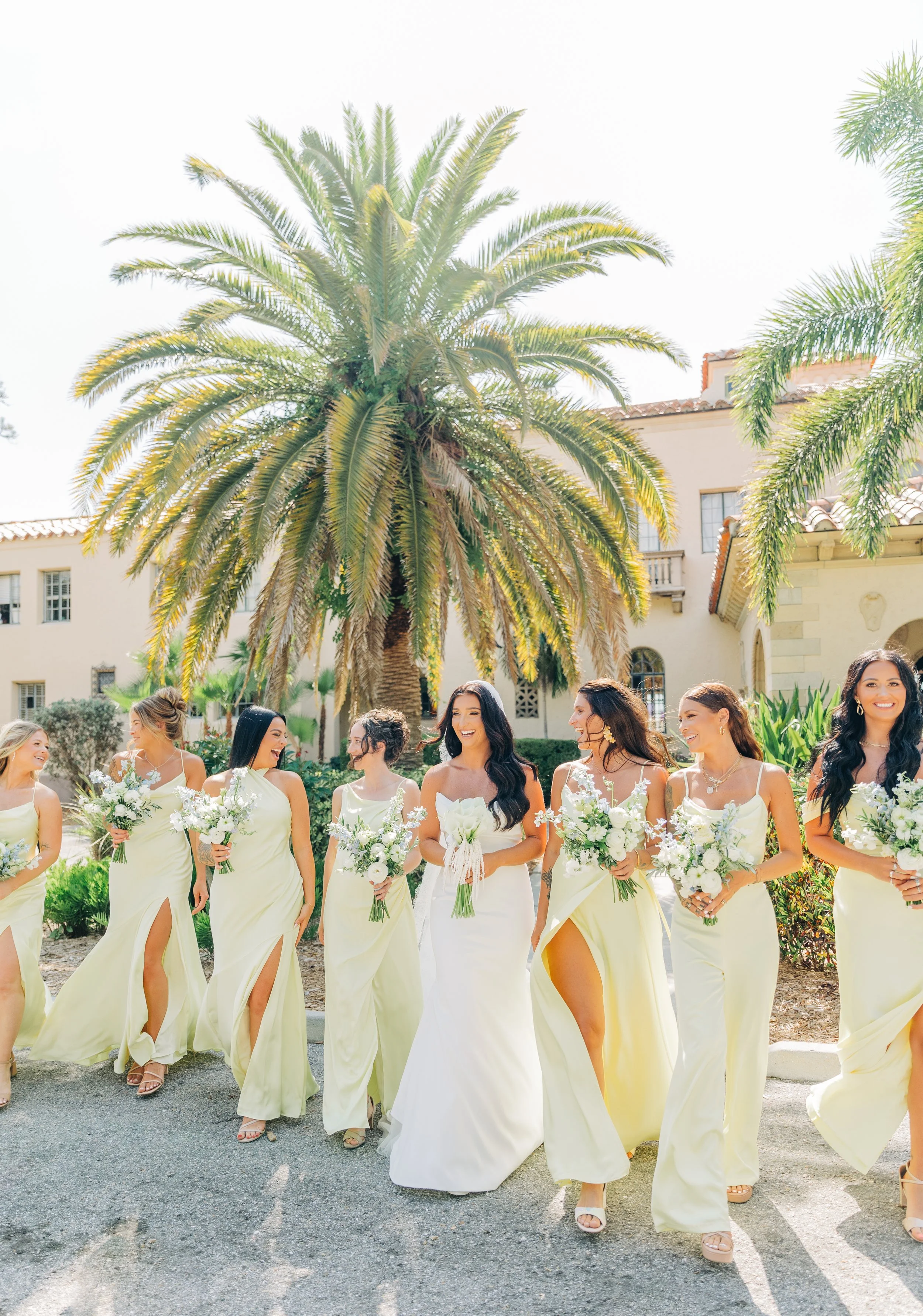 Group of women in yellow dresses holding bouquets of white flowers, walking outdoors on a sunny day, with palm trees and a building in the background.