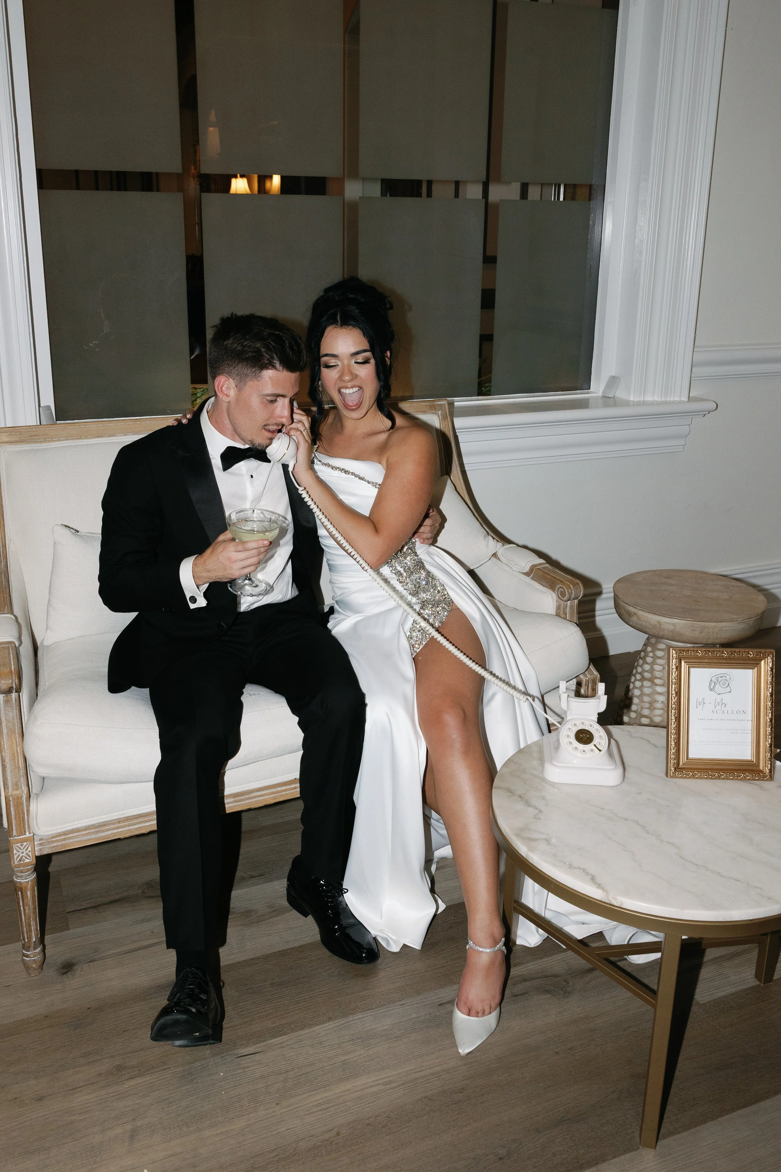 A man in a tuxedo and a woman in a wedding dress sitting on a cream-colored sofa, talking on a rotary phone, with the woman holding a cocktail glass. The woman is dressed in a wedding gown with high heel shoes. There is a marble-topped side table nex