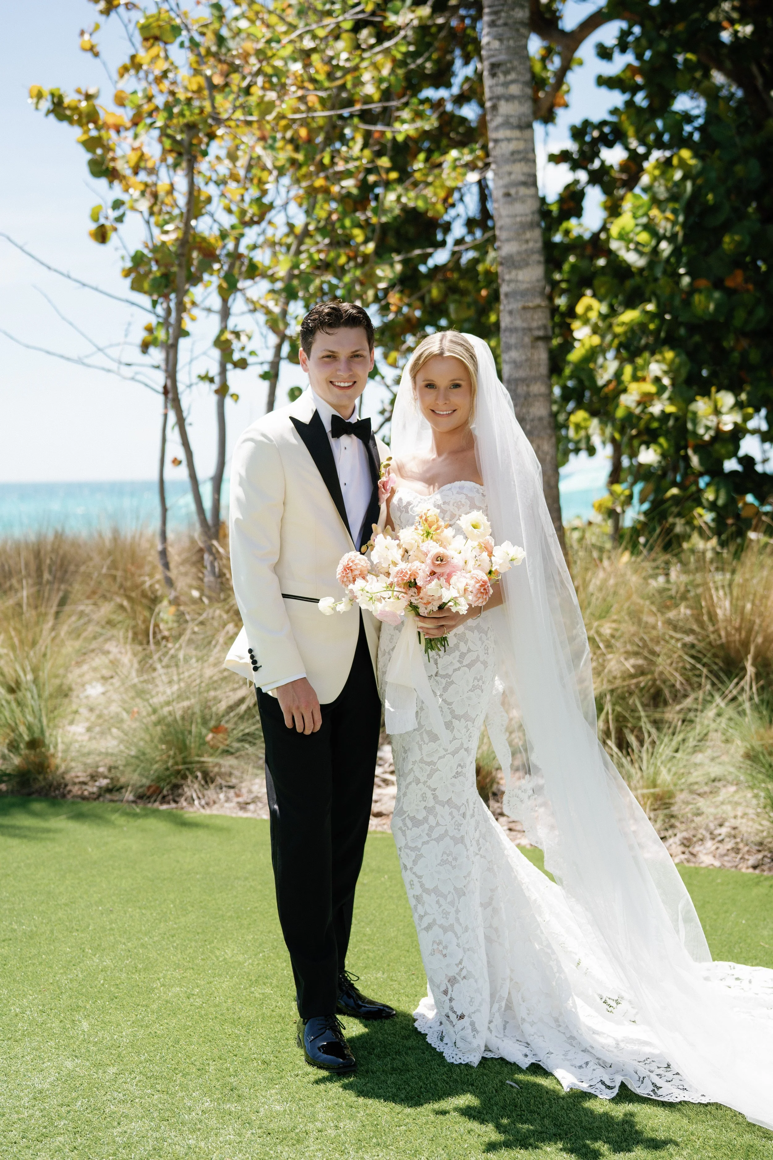 A bride and groom standing outdoors on a sunny day with trees and the beach in the background, smiling for the camera. The groom is wearing a white tuxedo jacket with black lapels and a black bow tie. The bride is wearing a white lace wedding gown an
