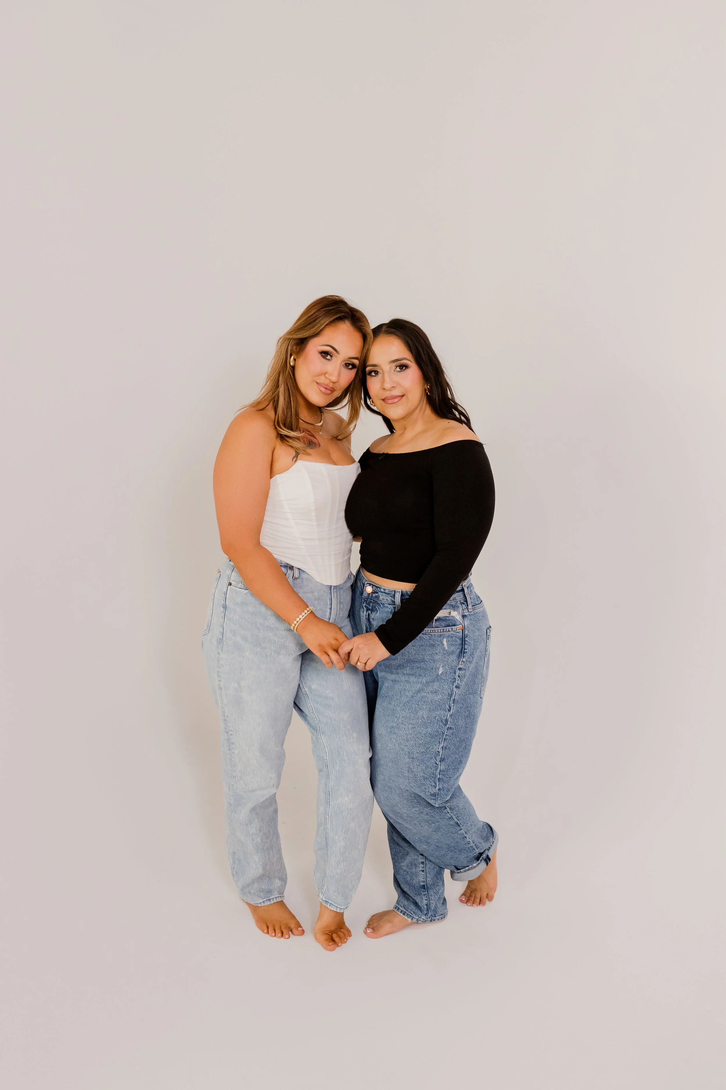 Two women standing close together, holding hands, barefoot, smiling, against a plain white background. One in a white strapless top and light jeans, the other in a black long-sleeve top and light jeans.