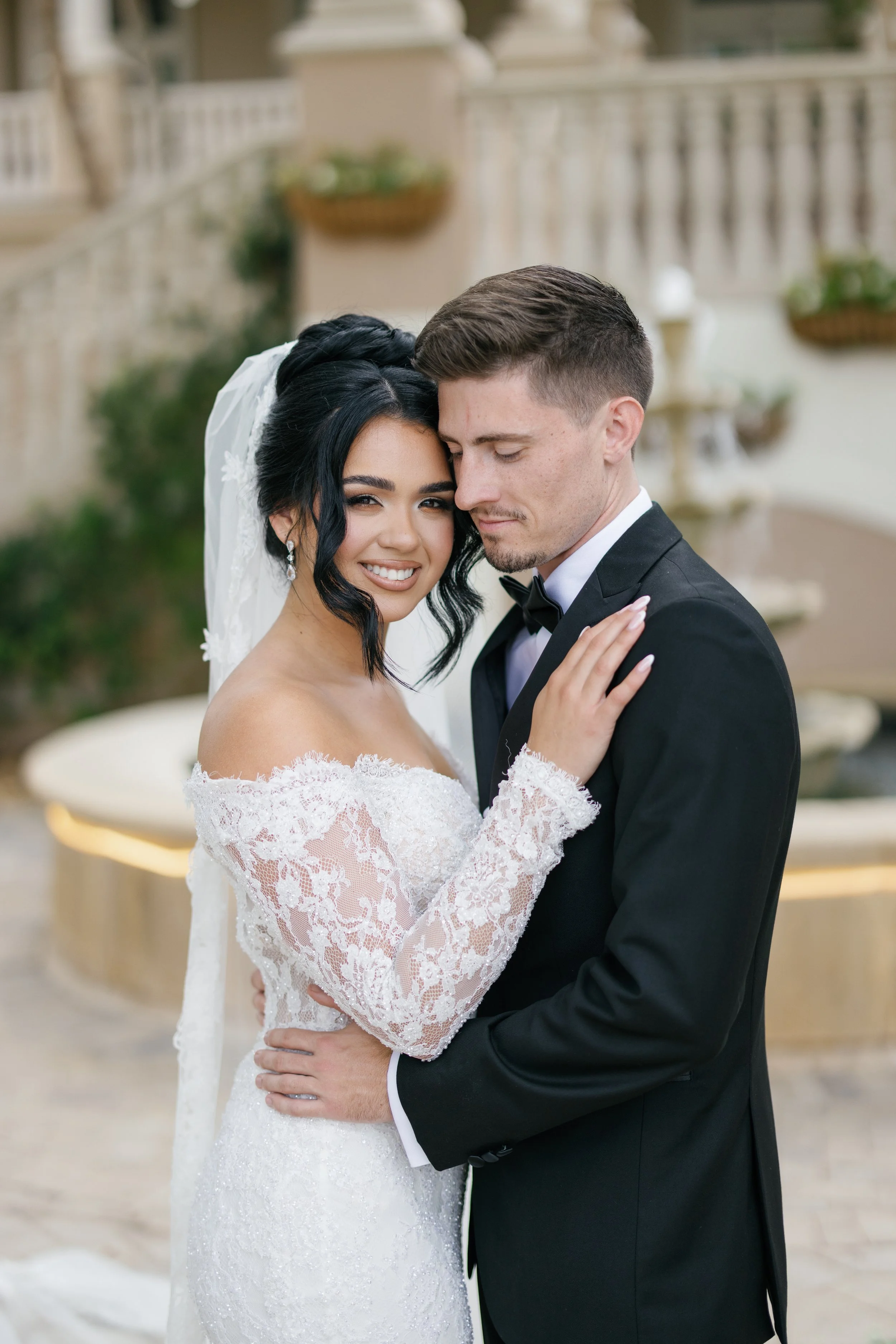 A bride and groom are standing close together, with the bride smiling at the camera and the groom with his eyes closed, at an outdoor wedding. The bride is wearing a white lace off-shoulder wedding dress with lace sleeves and a veil, while the groom 