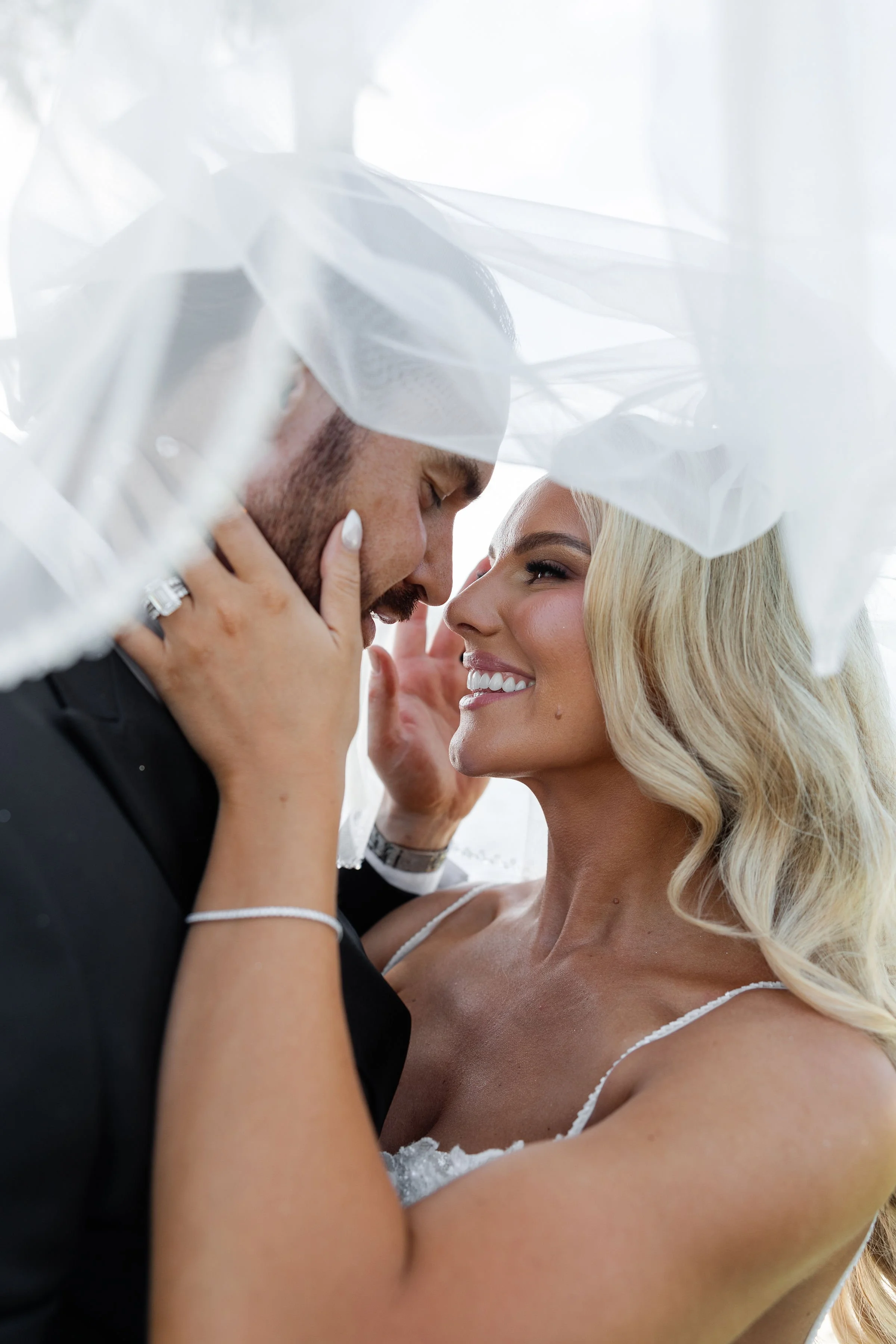 A couple, a man and a woman, are close together under a sheer white curtain. The man is wearing a black suit and the woman is wearing a white dress. They are touching foreheads and smiling at each other, with the woman holding the man's face gently.