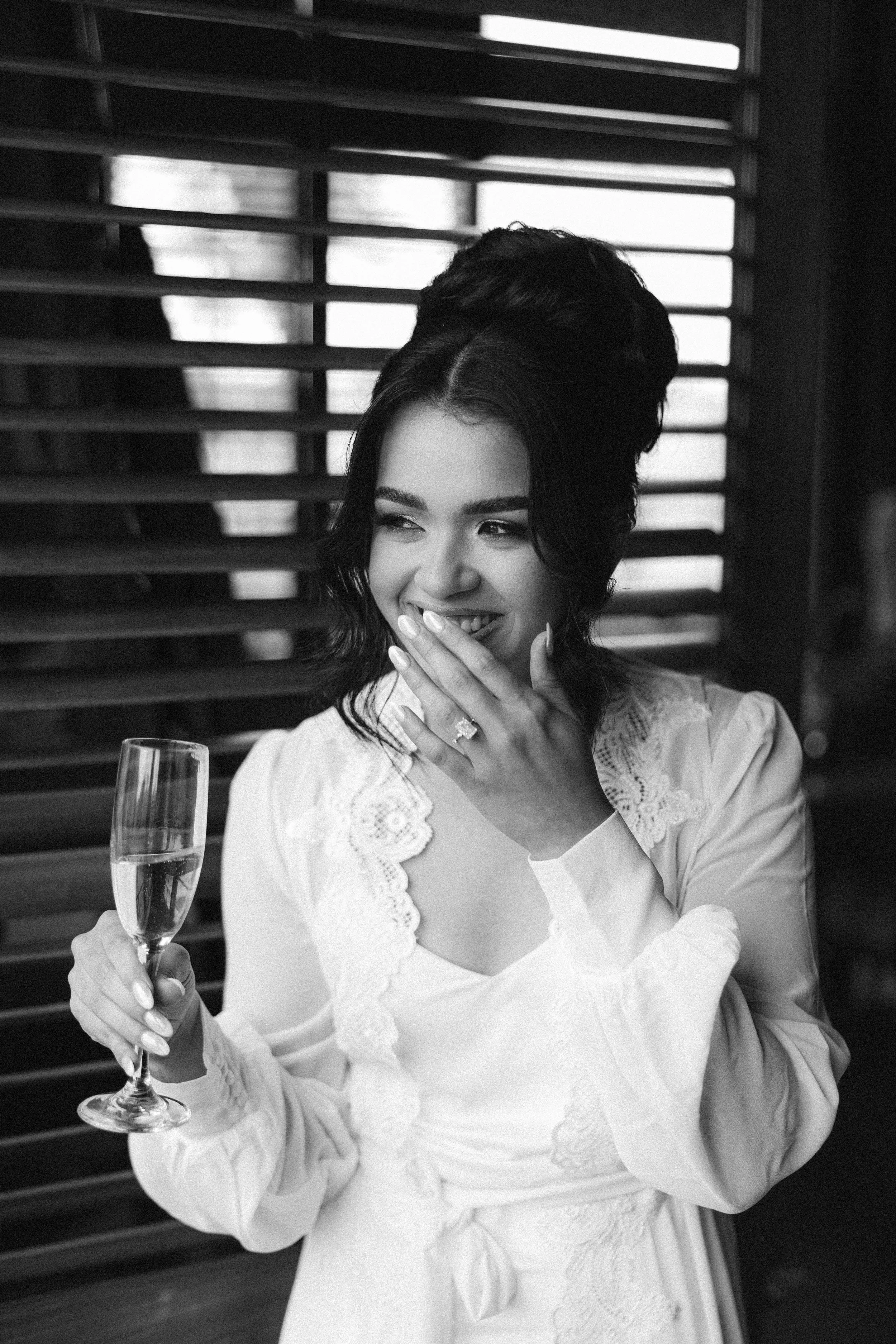A woman with dark, styled hair, wearing a light-colored embroidered dress, is smiling and covering her mouth with her hand while holding a glass of champagne. There are wooden blinds in the background.