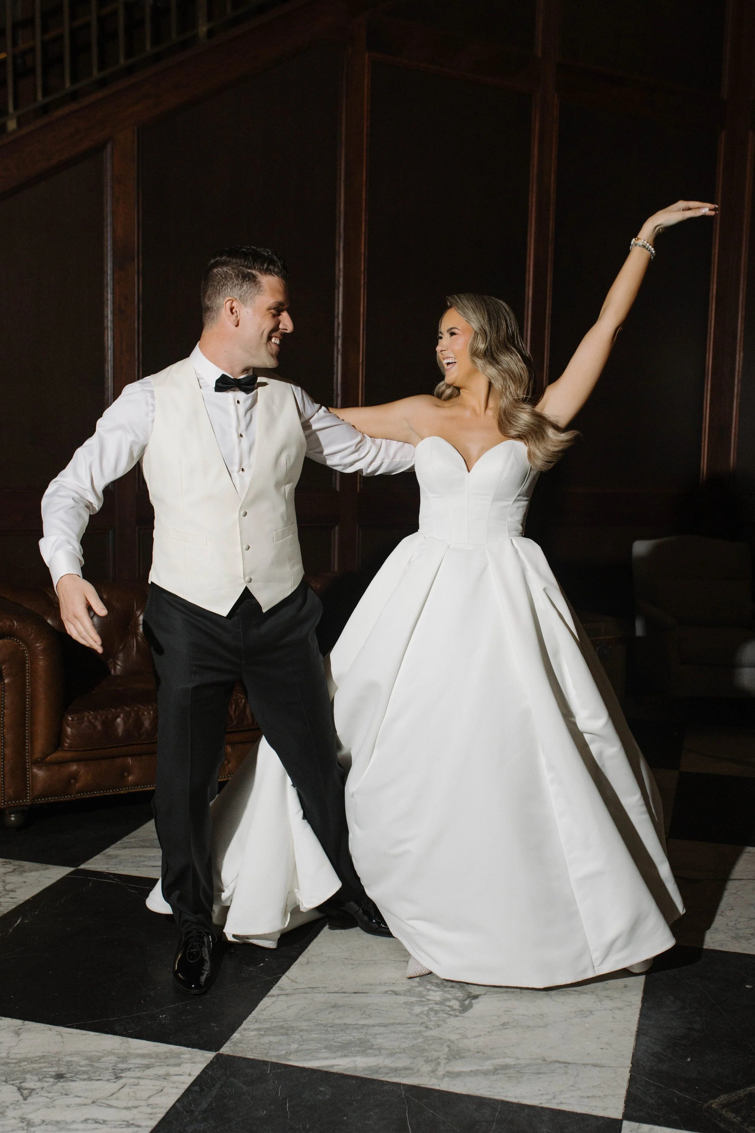 A bride and groom dancing together indoors. The groom is wearing a tuxedo vest with a bow tie, and the bride is in a white wedding gown with a full skirt and has long, wavy hair. They are smiling and looking at each other, with the bride raising one 