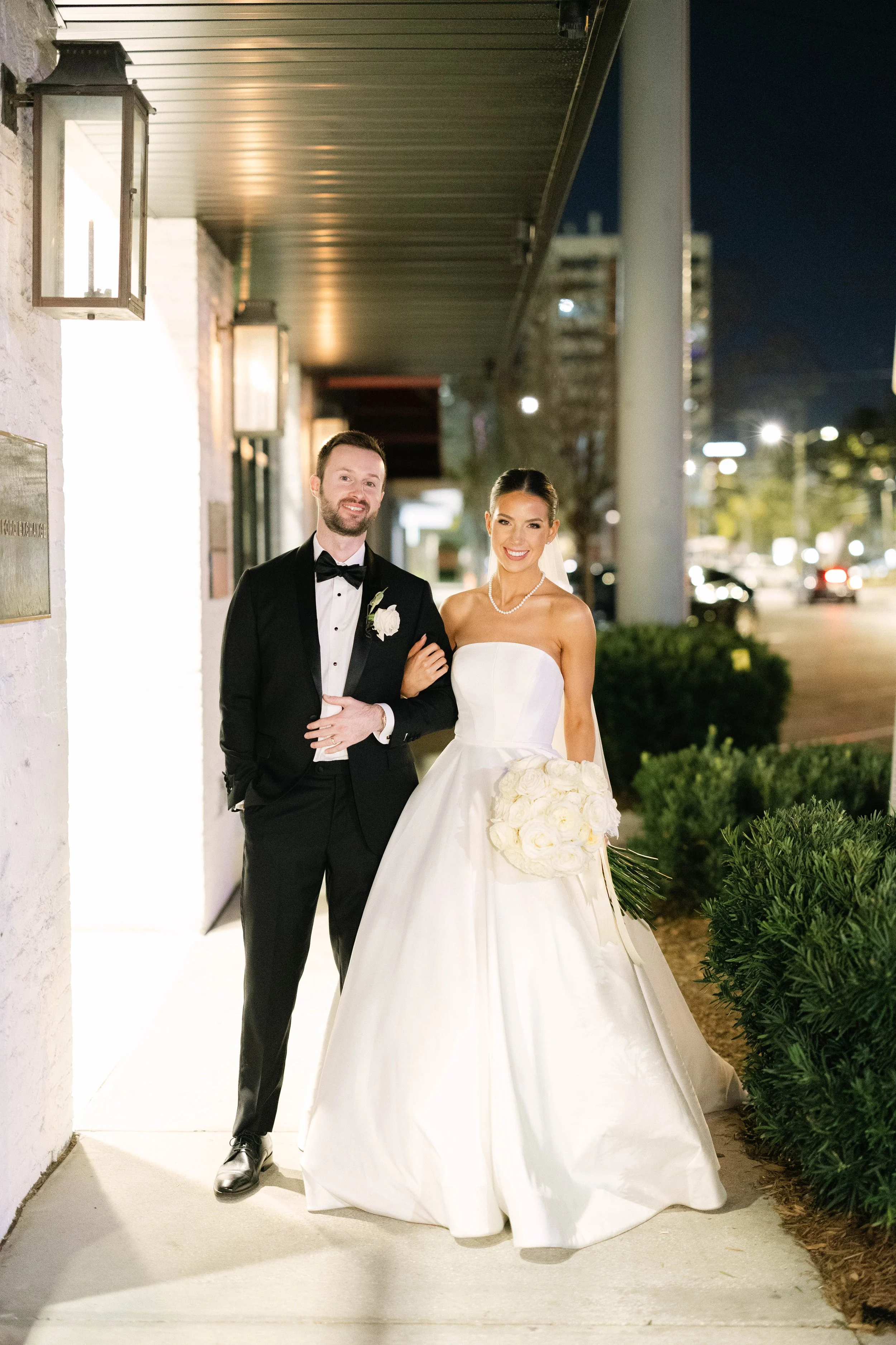 A newlywed couple stands outside at night. The groom wears a black tuxedo with a bow tie and boutonniere, and the bride wears a strapless white wedding gown with a pearl necklace, holding a bouquet of white roses. They are smiling and standing arm in