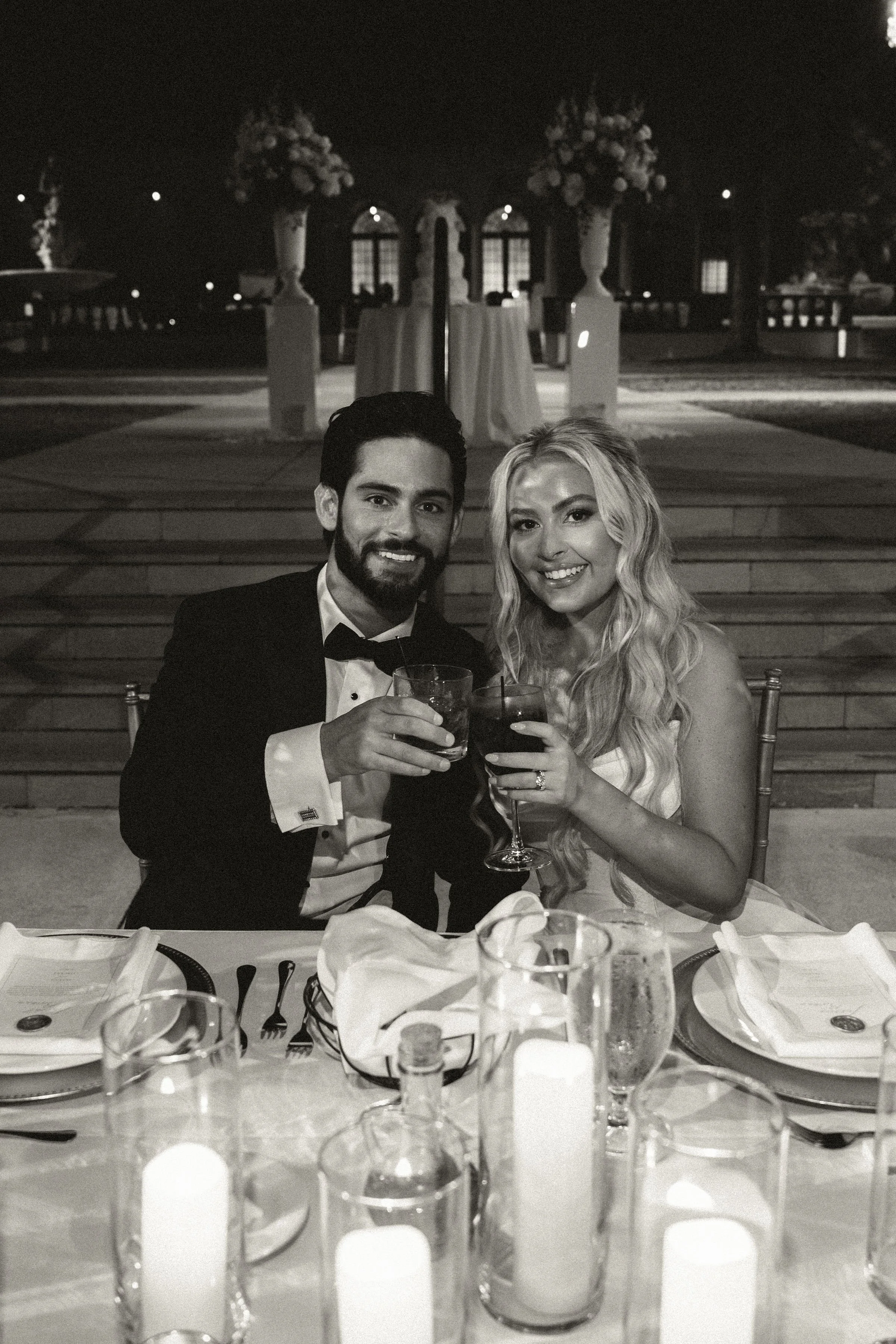 A couple dressed in formal attire celebrating at a dinner table with candles, glasses, and plates, in an outdoor setting at night.