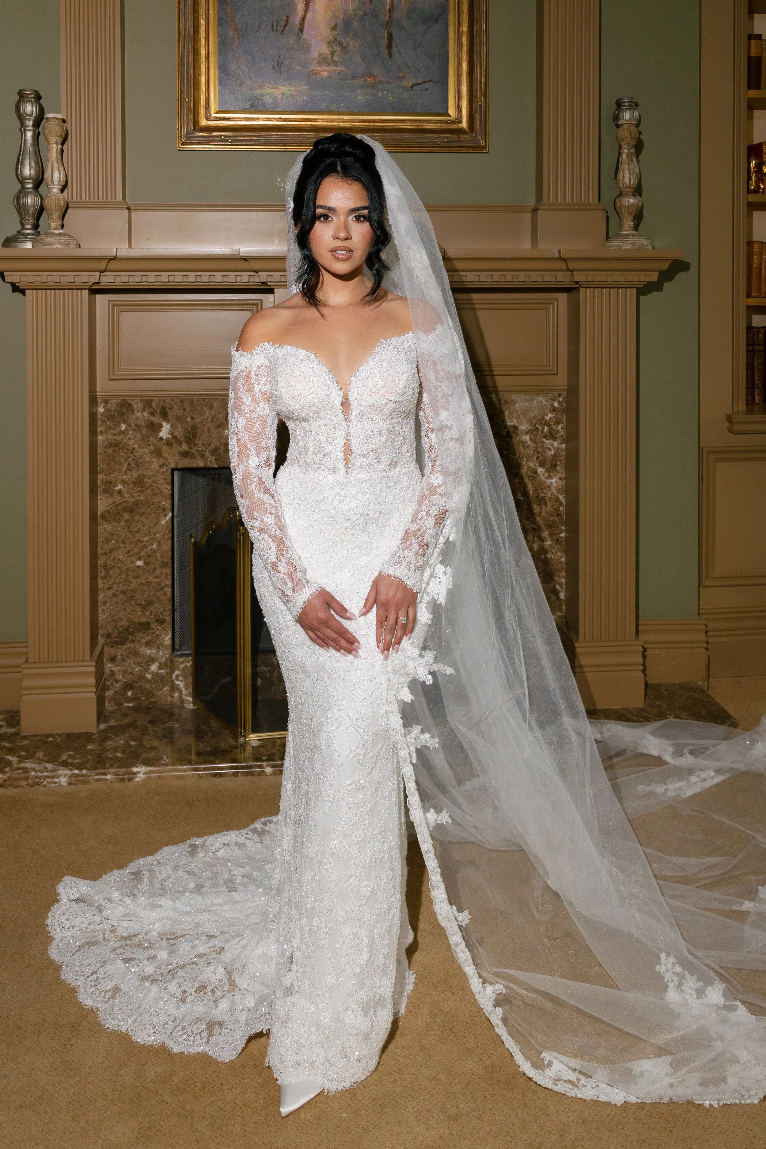 A woman in a white lace wedding gown with long sleeves and a train, wearing a veil, standing in front of a fireplace in a room with green walls and bookcases.