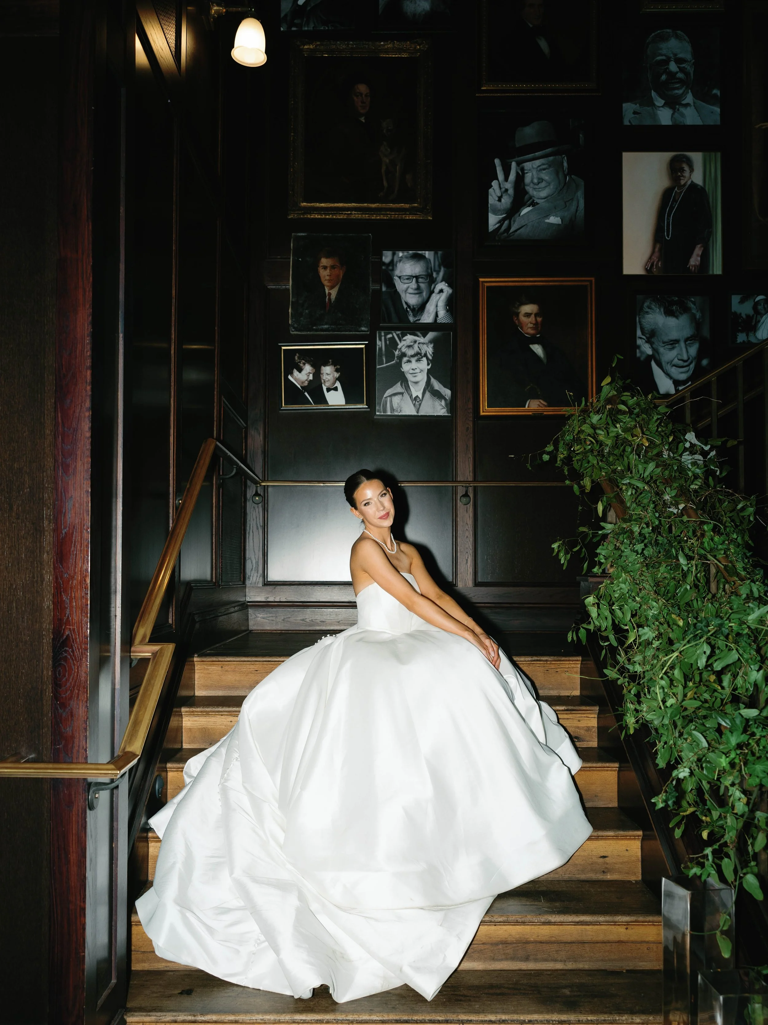 A woman in a white wedding dress sitting on a staircase in a dimly lit room with dark wooden walls. The wall behind her is decorated with framed photographs and paintings of various people, some in black and white and others in color.