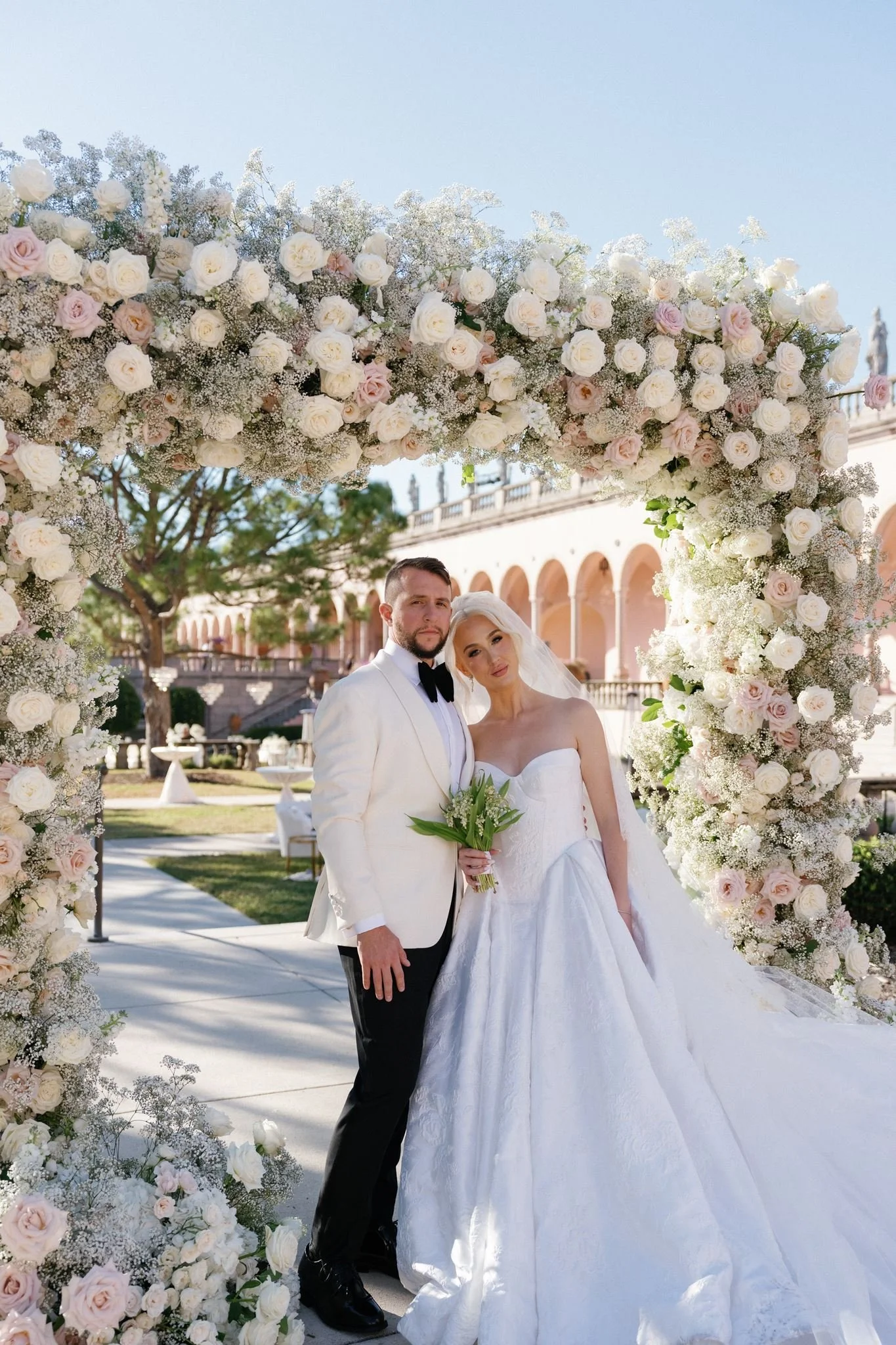 A newlywed couple in wedding attire standing under a floral arch made of white and light pink roses and baby's breath, outdoors on a bright sunny day with a building with arches in the background.