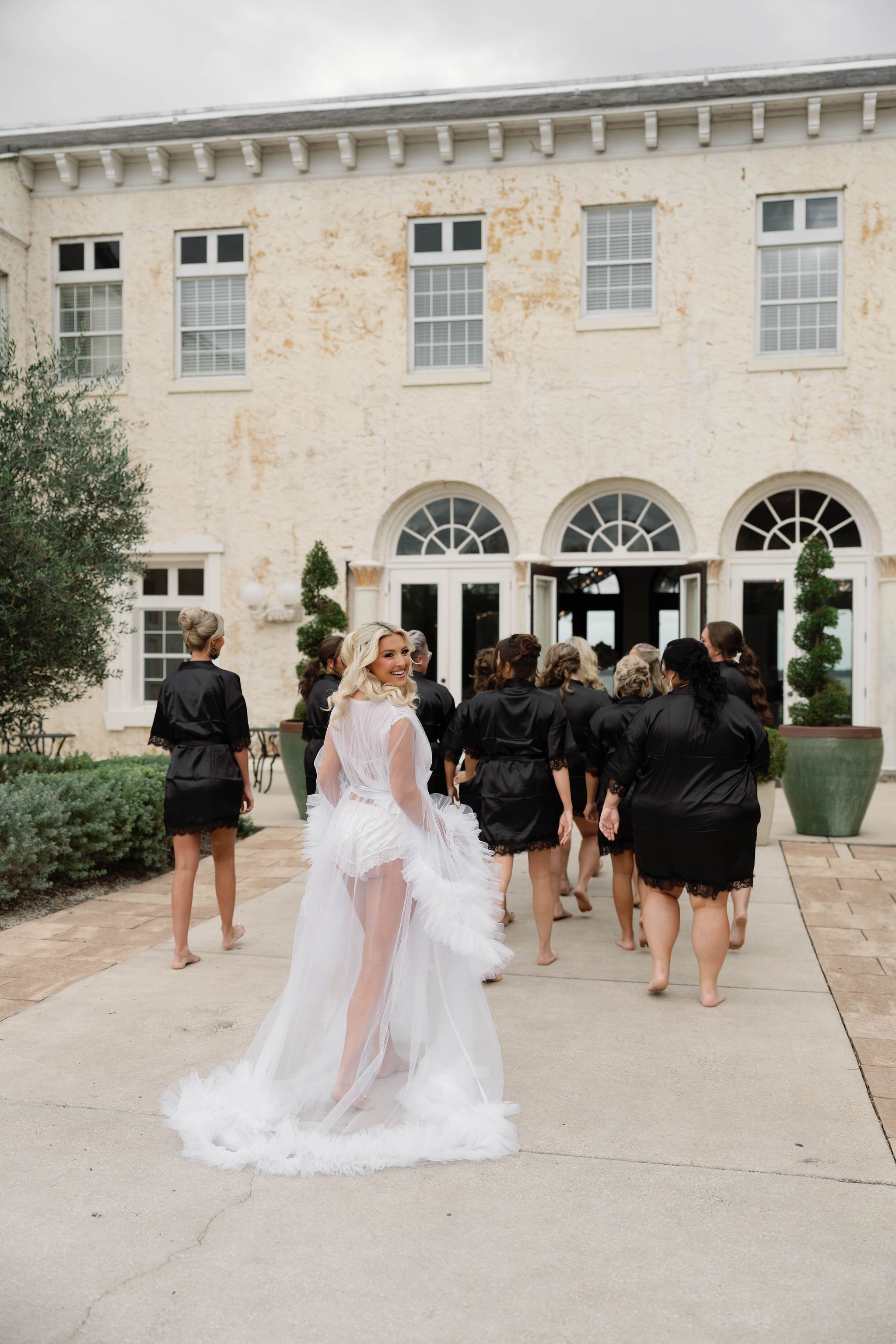 A bride in a white wedding dress with a sheer overlay and ruffled train standing outside in front of a group of women in black robes, with a large stone building with multiple windows in the background.
