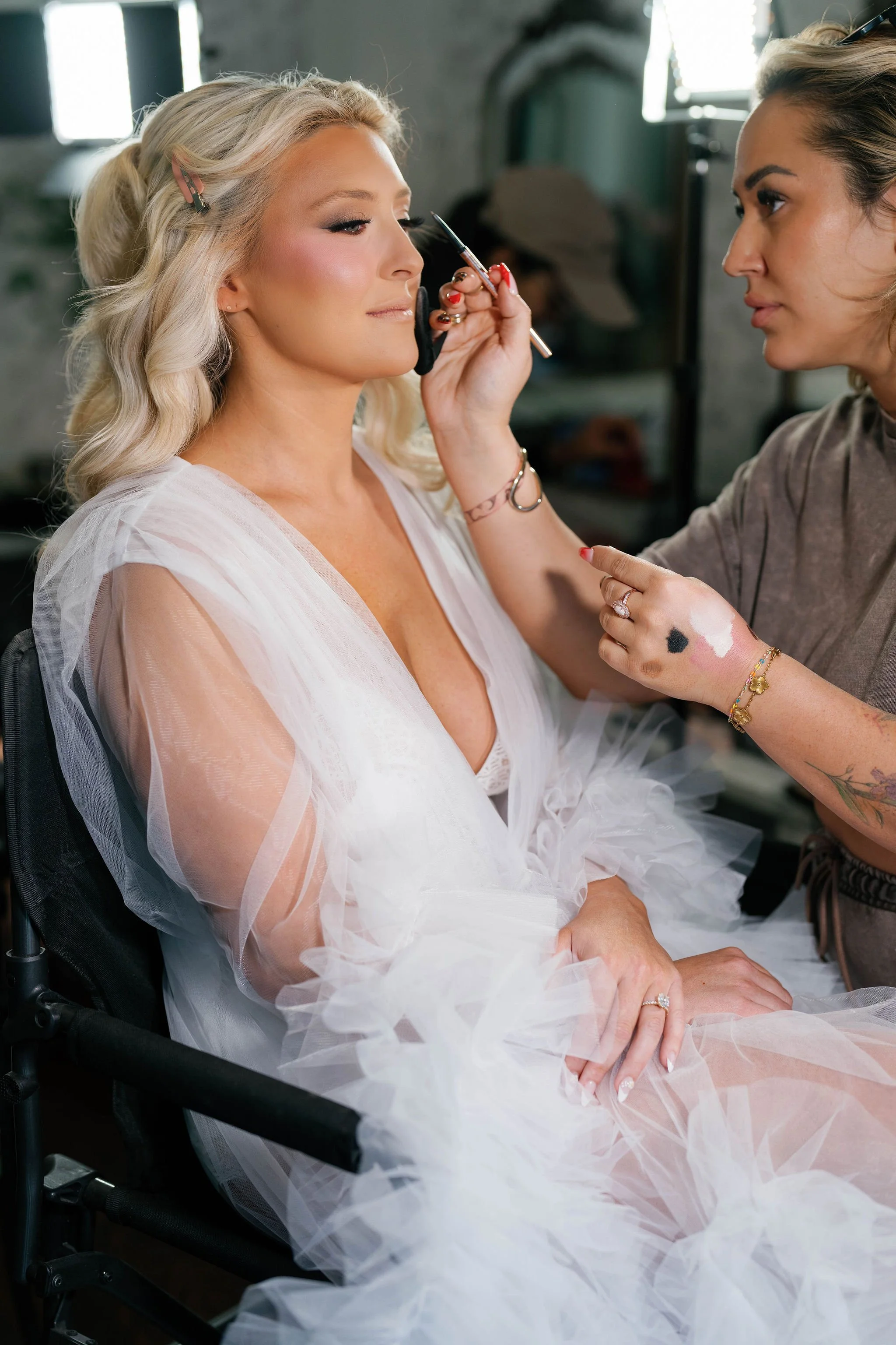 A woman with blonde hair in a white wedding dress sitting in a wheelchair as a makeup artist applies makeup to her face.