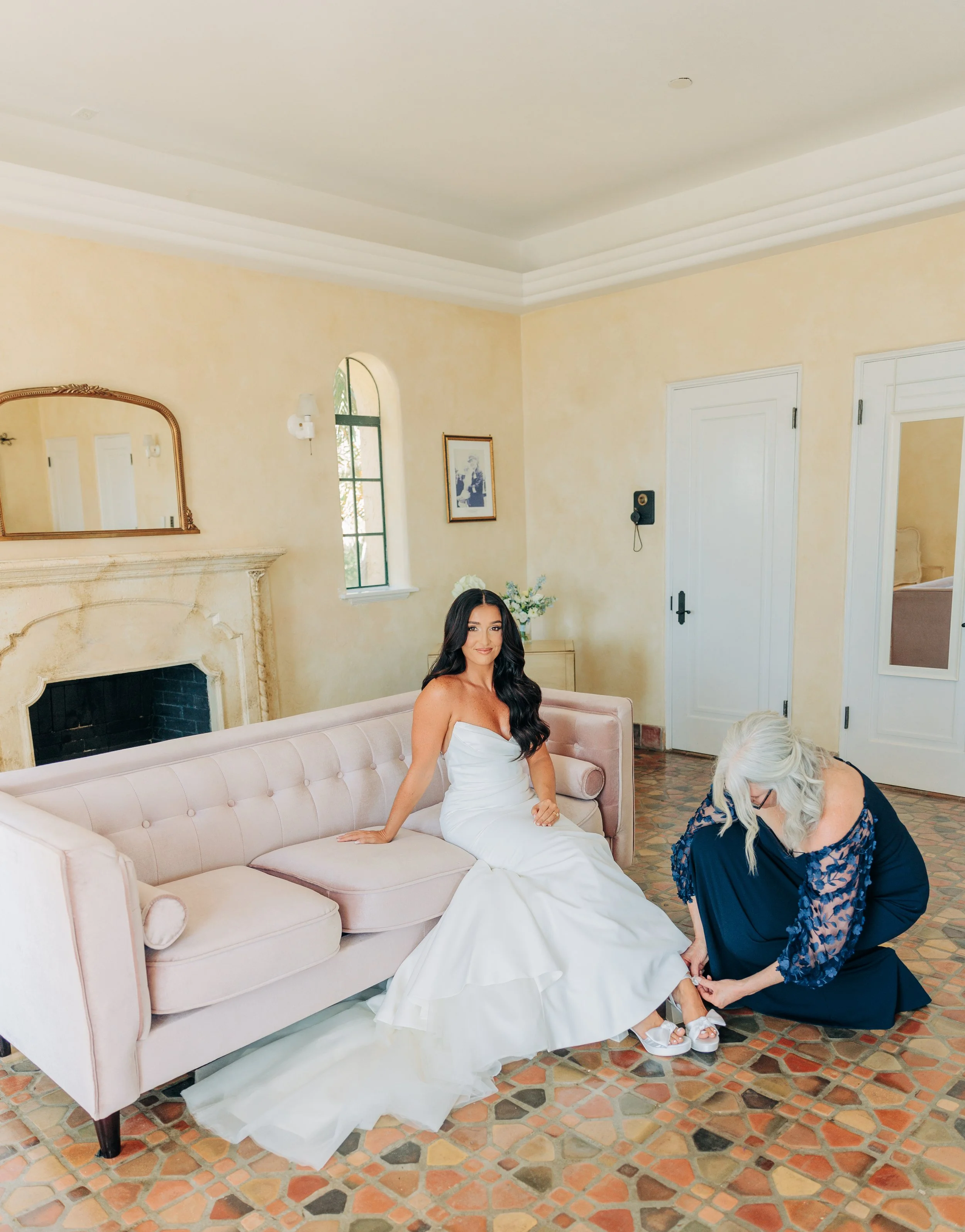 A woman in a white wedding dress sitting on a pink sofa while a woman in a dark dress helps her put on white shoes.