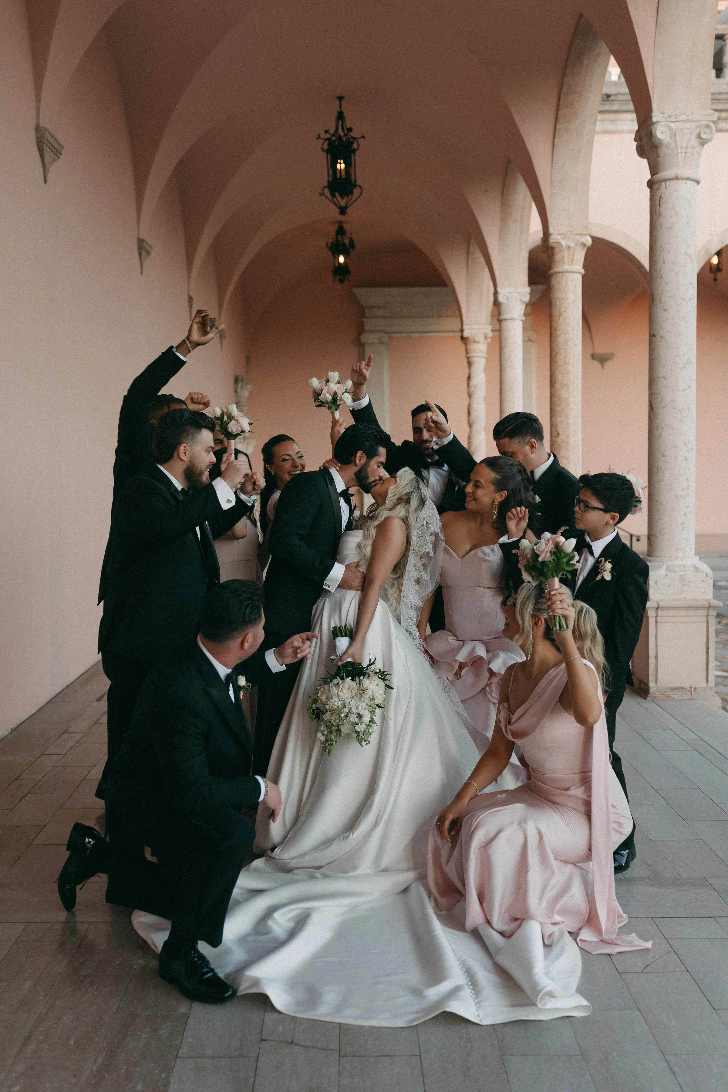 A wedding party standing in a covered outdoor corridor with pink arches and columns, celebrating and holding bouquets. The bride and groom are kissing, surrounded by bridesmaids and groomsmen.
