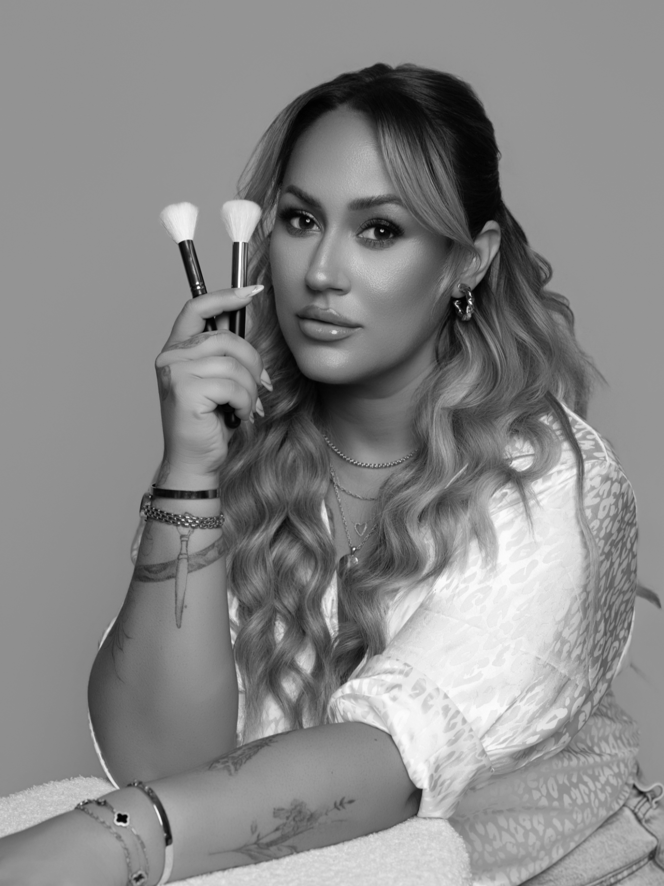 Black and white photo of a woman with long, wavy hair holding two makeup brushes, looking at the camera with a confident expression. She is wearing earrings, layered necklaces, and has tattoos and bracelets on her arm.