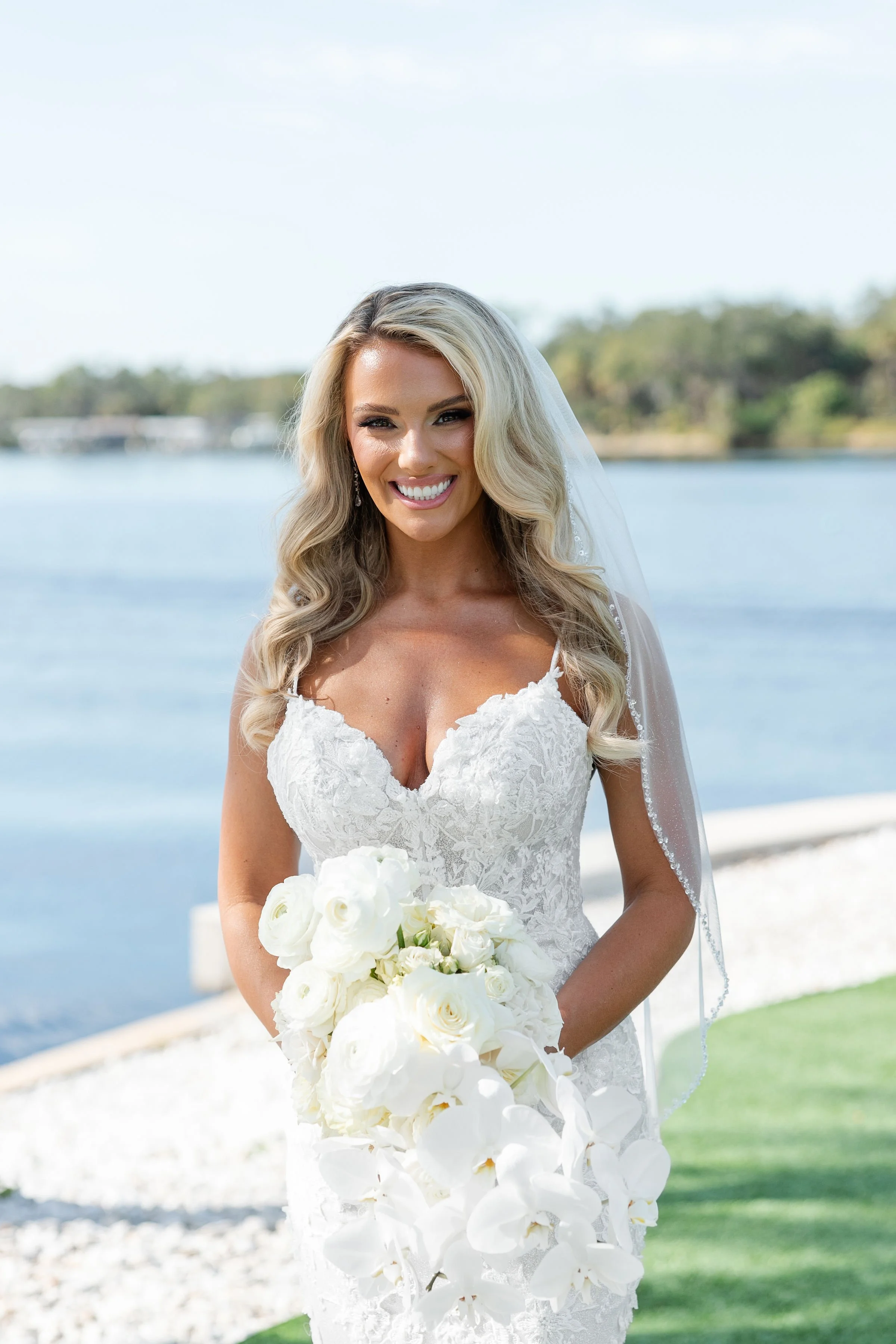 Bride in a white lace wedding dress holding a bouquet of white flowers, smiling outdoors near a body of water with trees in the background.