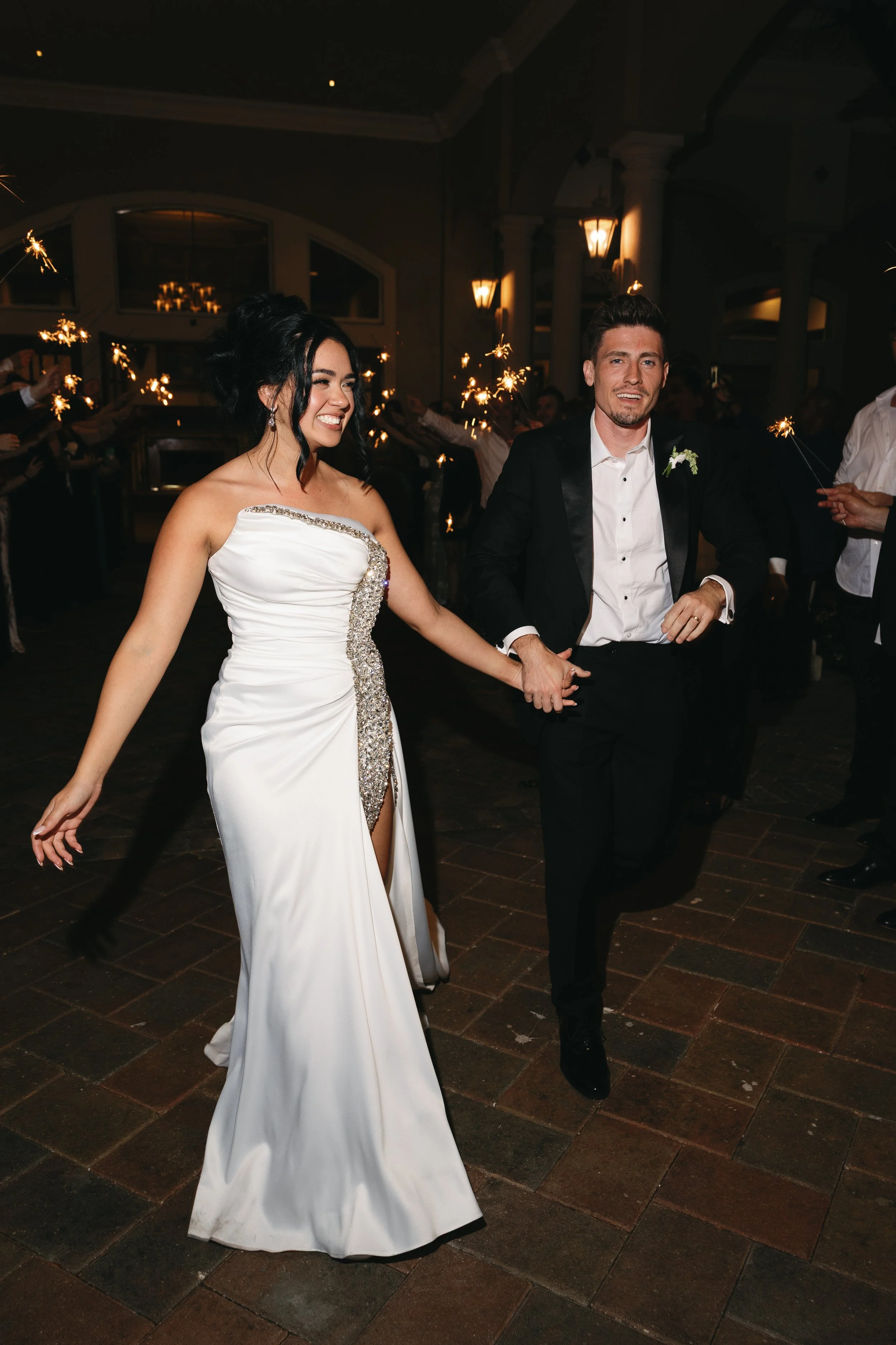 A bride and groom smiling and holding hands on the dance floor during their wedding celebration, with guests holding sparklers in the background.