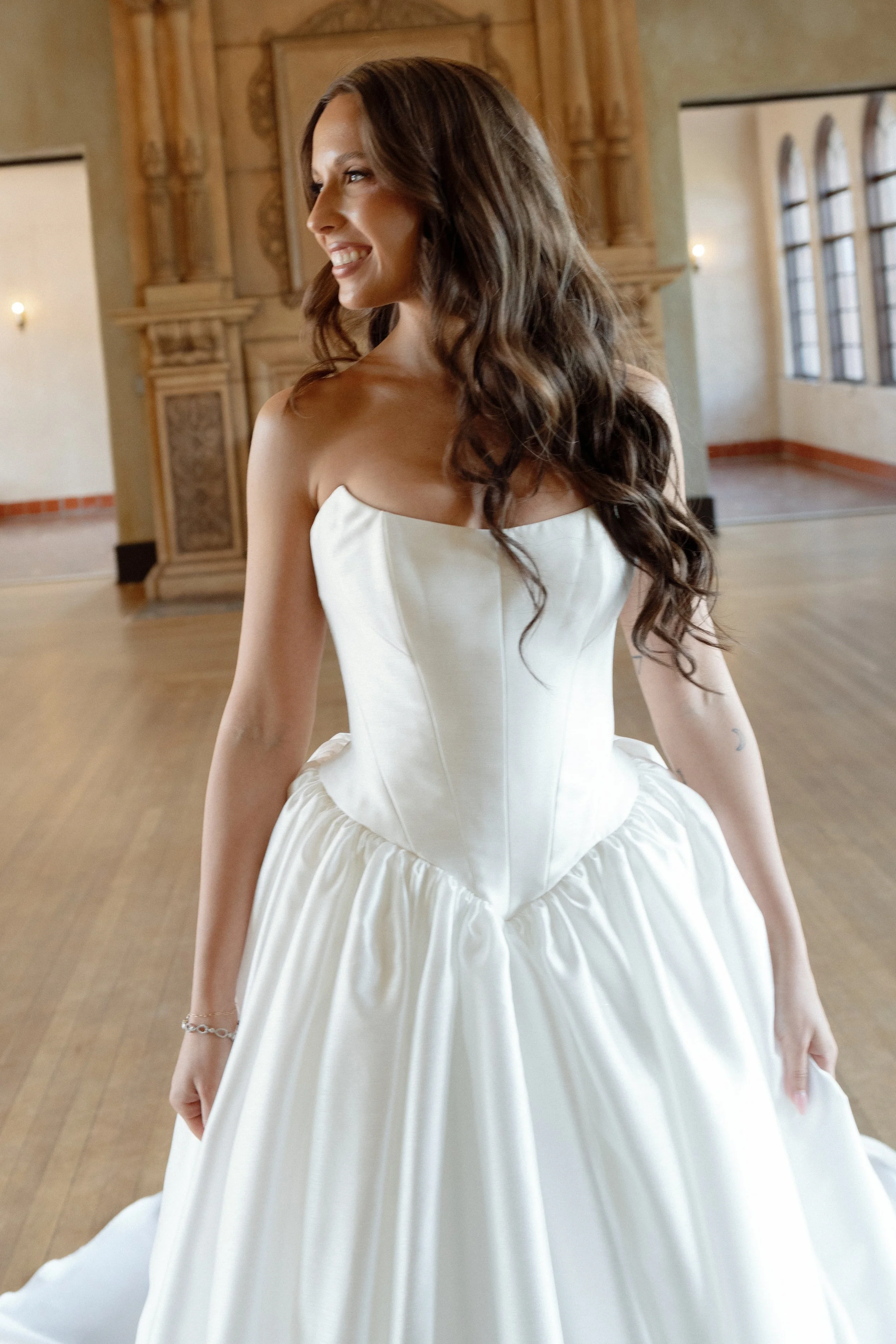 A smiling woman in a white wedding dress standing in a spacious, well-lit room with large windows and wooden floors.