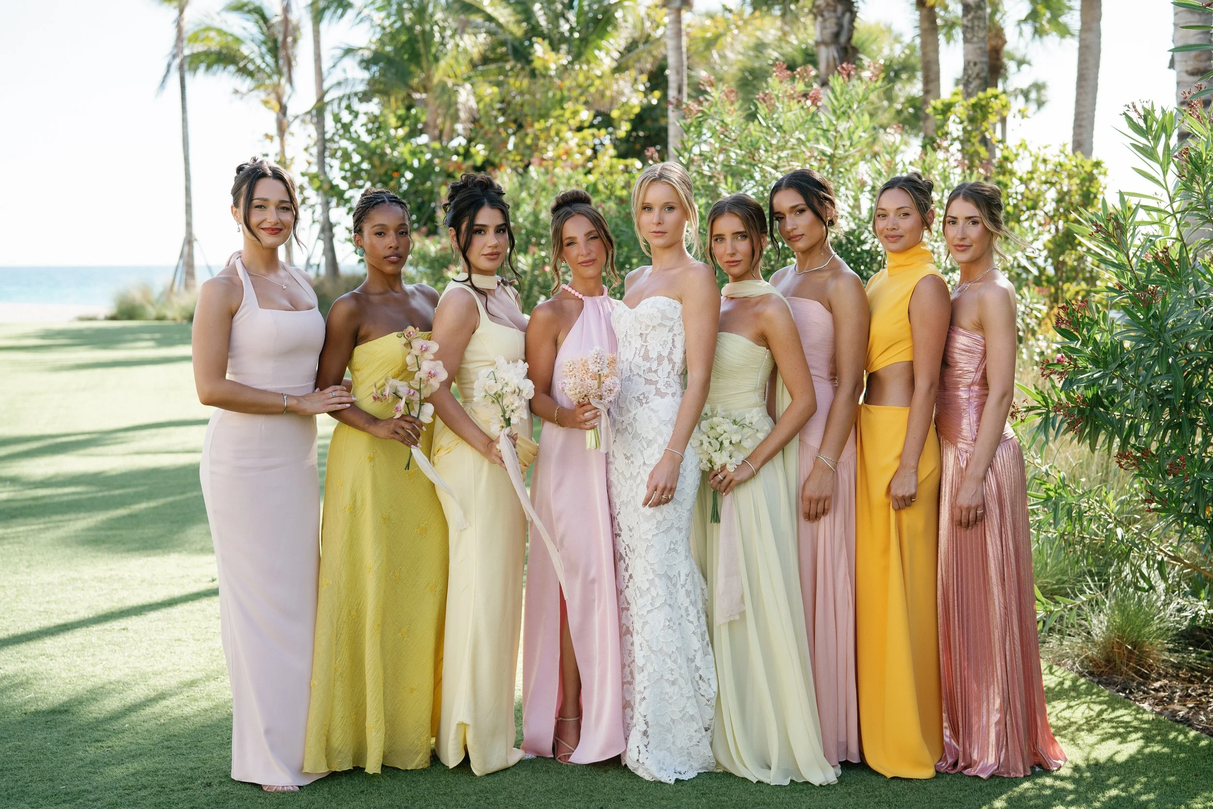 A group of eleven women in colorful dresses standing outdoors, with trees and a beach in the background, on a sunny day.