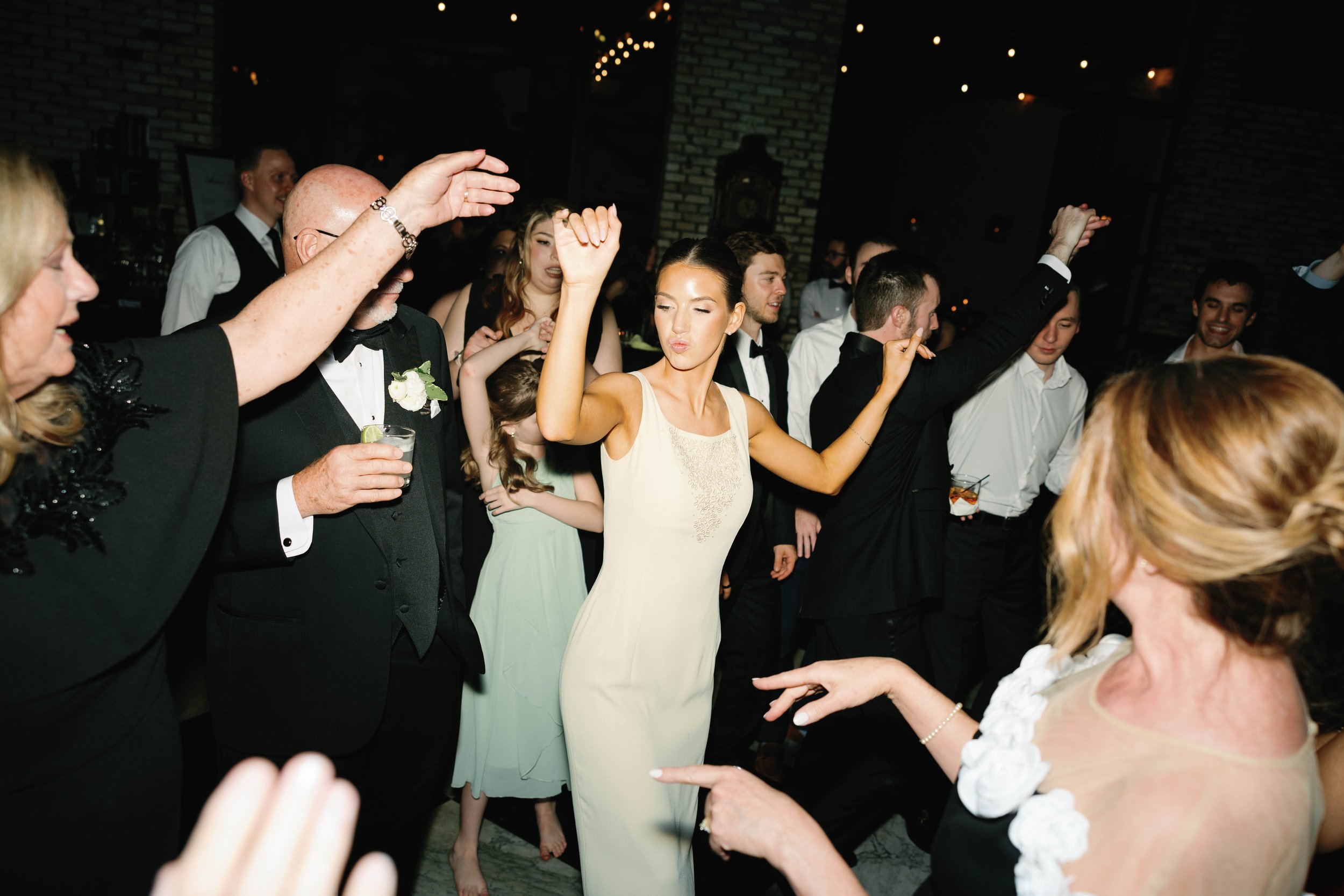Group of people dancing and celebrating at a party, dressed in formal attire with some holding drinks.