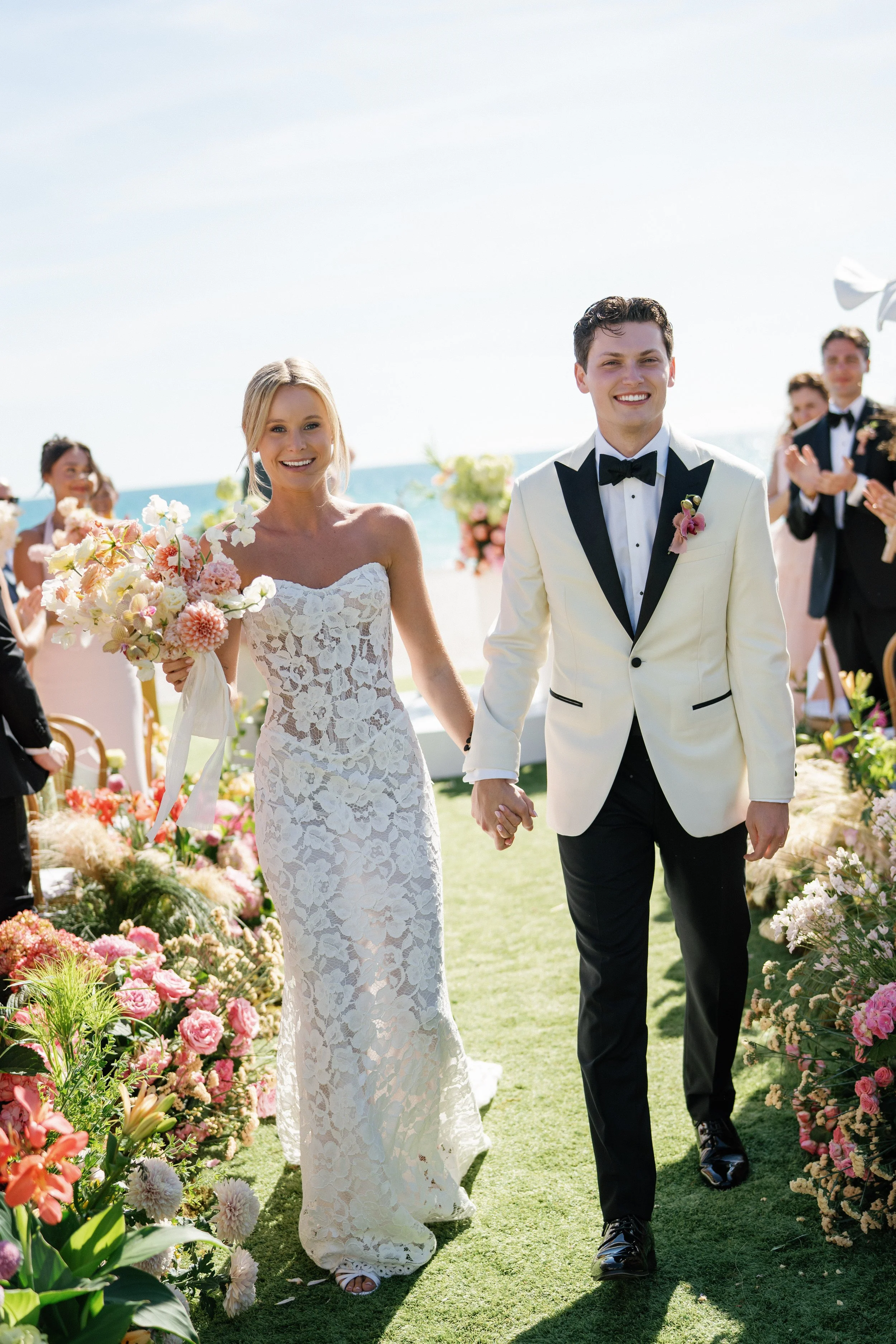 A bride and groom holding hands, walking down a flower-filled aisle outdoors by the beach, smiling happily.