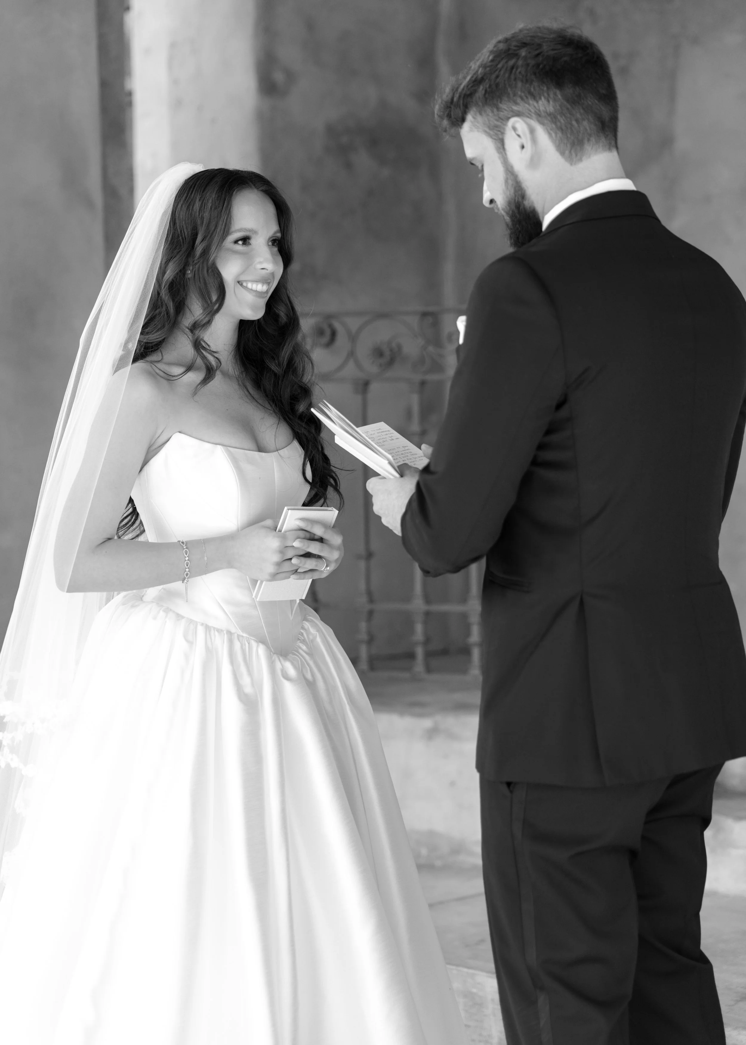 A black-and-white photograph of a bride and groom exchanging wedding vows inside a building with stone walls. The bride is smiling, wearing a strapless wedding dress with a veil, holding a small box, and a bracelet on her wrist. The groom, facing the bride, is in a dark suit with a white shirt and is holding a piece of paper or vows. They are standing close, with a decorative metal railing in the background.