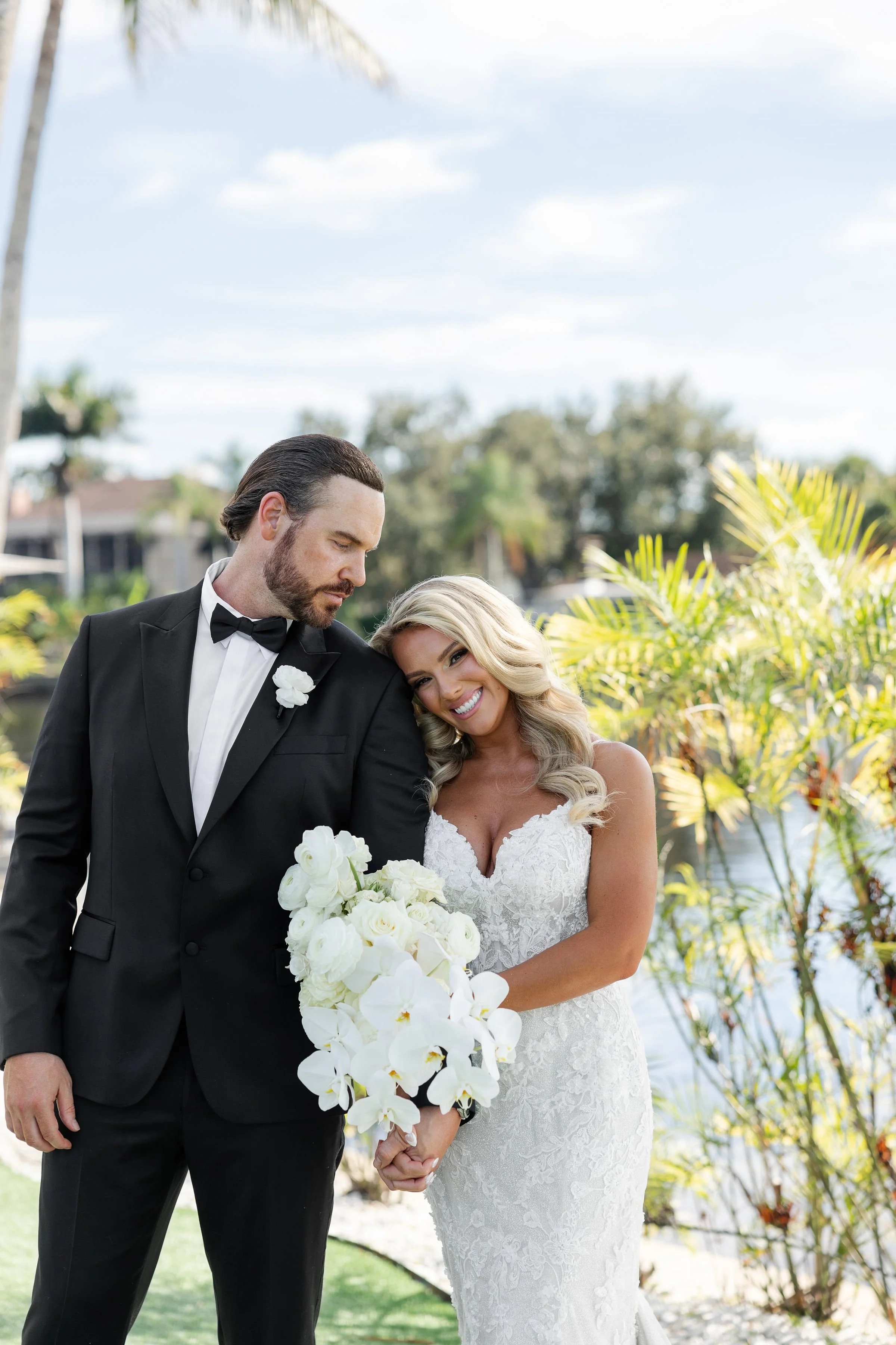 A bride and groom standing outside by a water feature, holding hands, with the bride smiling and the groom looking down, surrounded by lush greenery and palm trees.