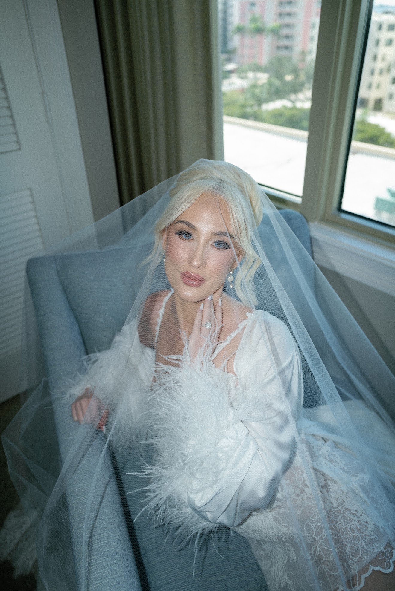 A woman with blonde hair styled in an updo, wearing a wedding dress with lace and feather details, sitting on a gray chair near a large window.