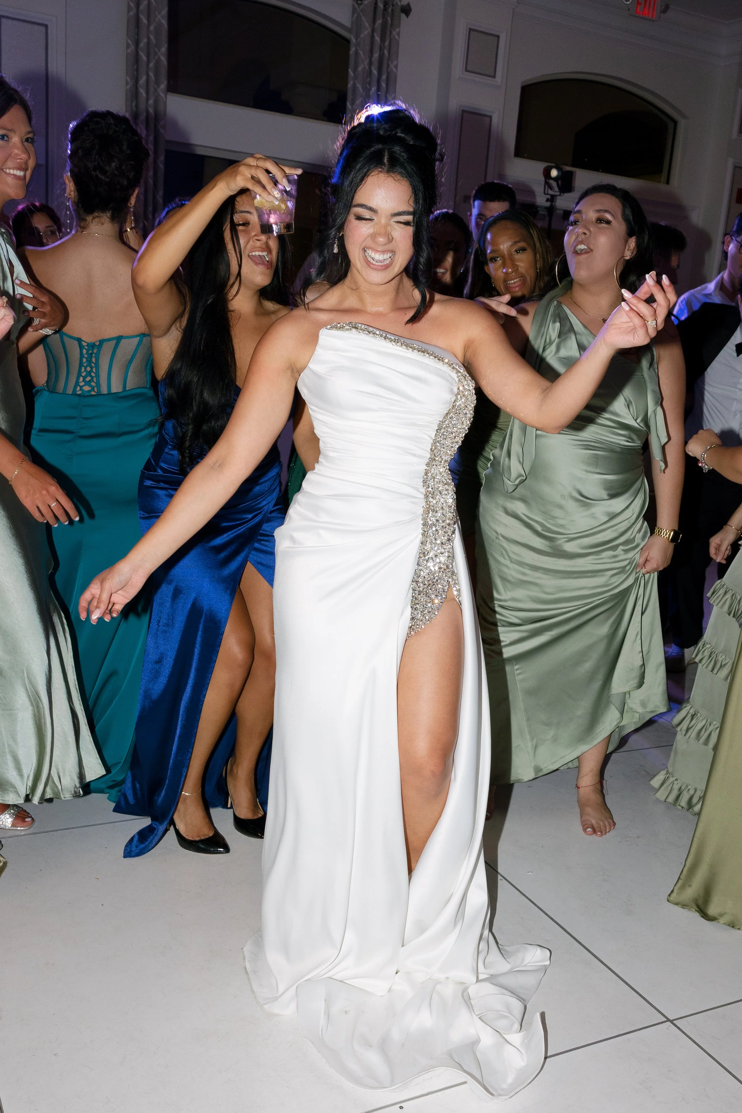 A woman in a white gown dancing at a celebration, surrounded by other women in colorful dresses.